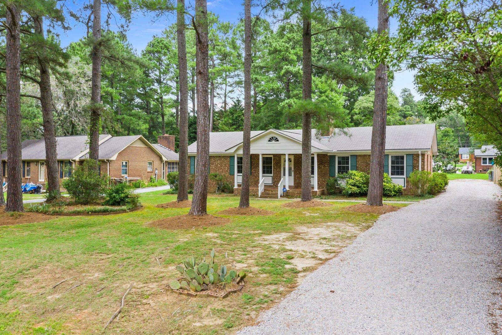 Single-story residential home with a centered front door, flanked by windows, and a small front porch, set back from a gravel pathway in a neighborhood with tall pine trees and green lawns.