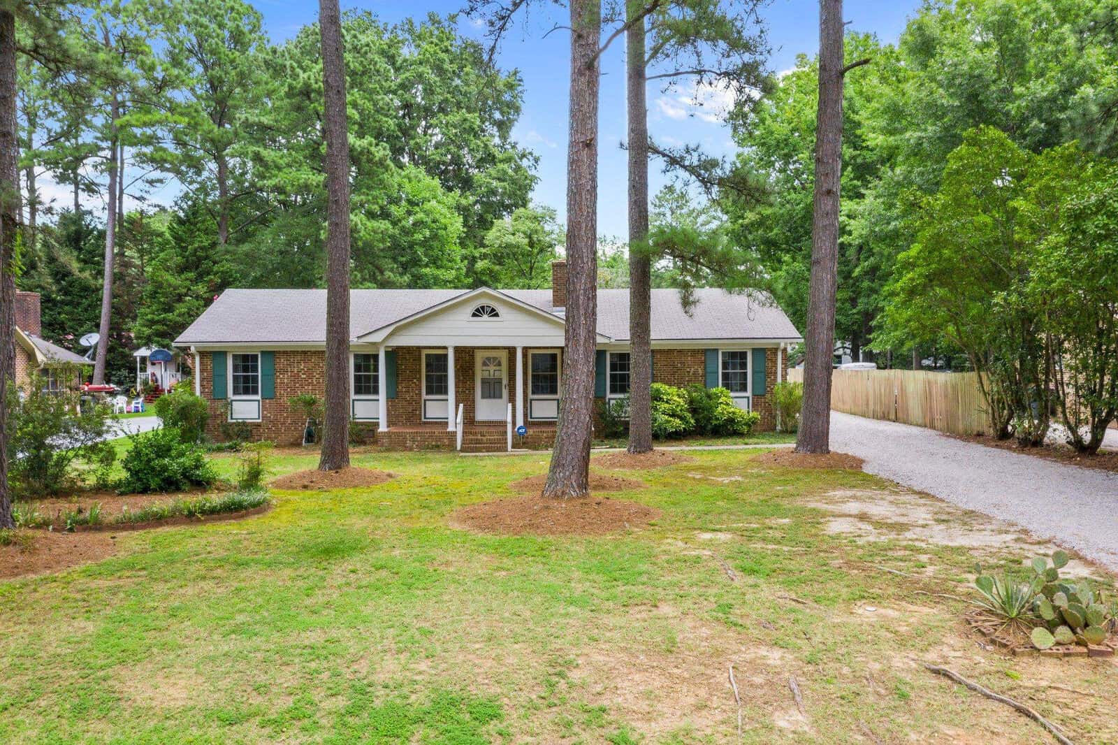 A single-story residential home with a brick facade, white trim, and a centered front door, set back from a gravel driveway and surrounded by tall pine trees and a well-maintained lawn. a wooden privacy fence is visible on the right side of the property.