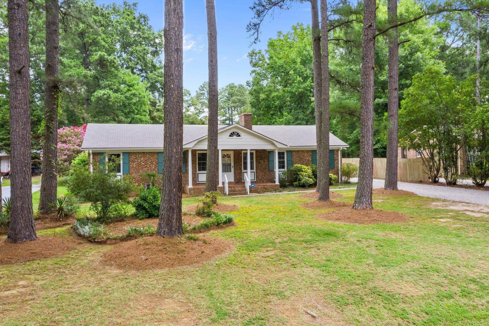 A single-story brick house with white trim, surrounded by tall pine trees and a well-manicured lawn, in a suburban setting.