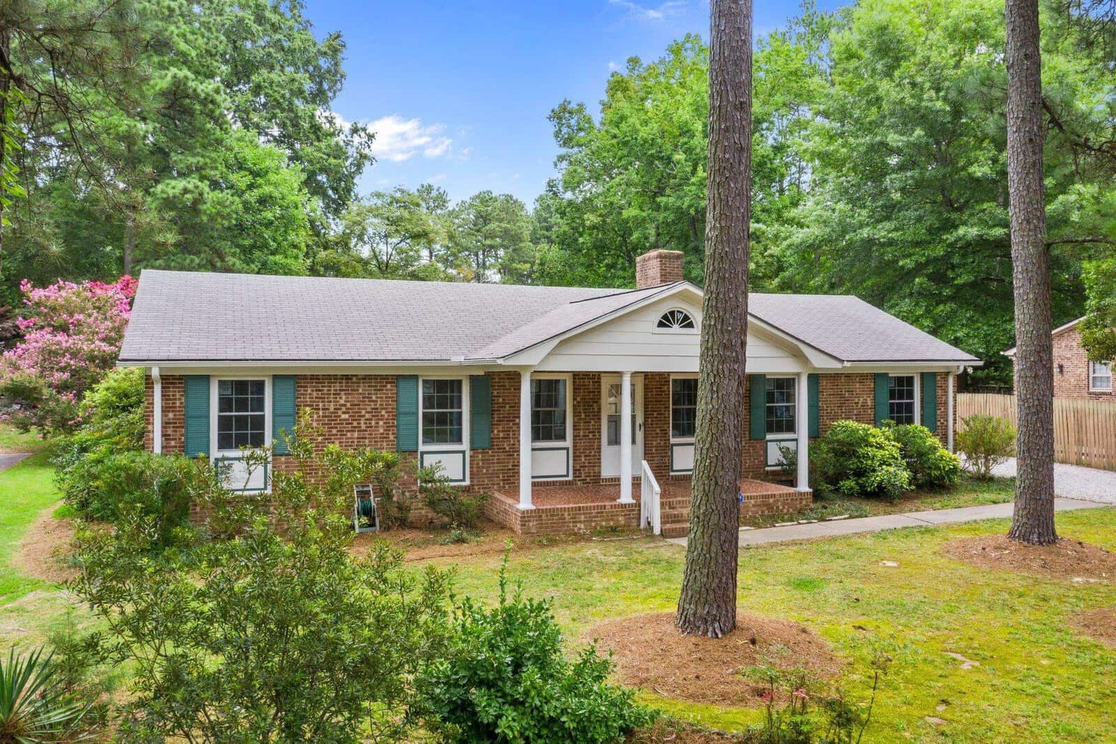 Single-story brick house with a white front door, flanked by two large pine trees and a neatly kept lawn.