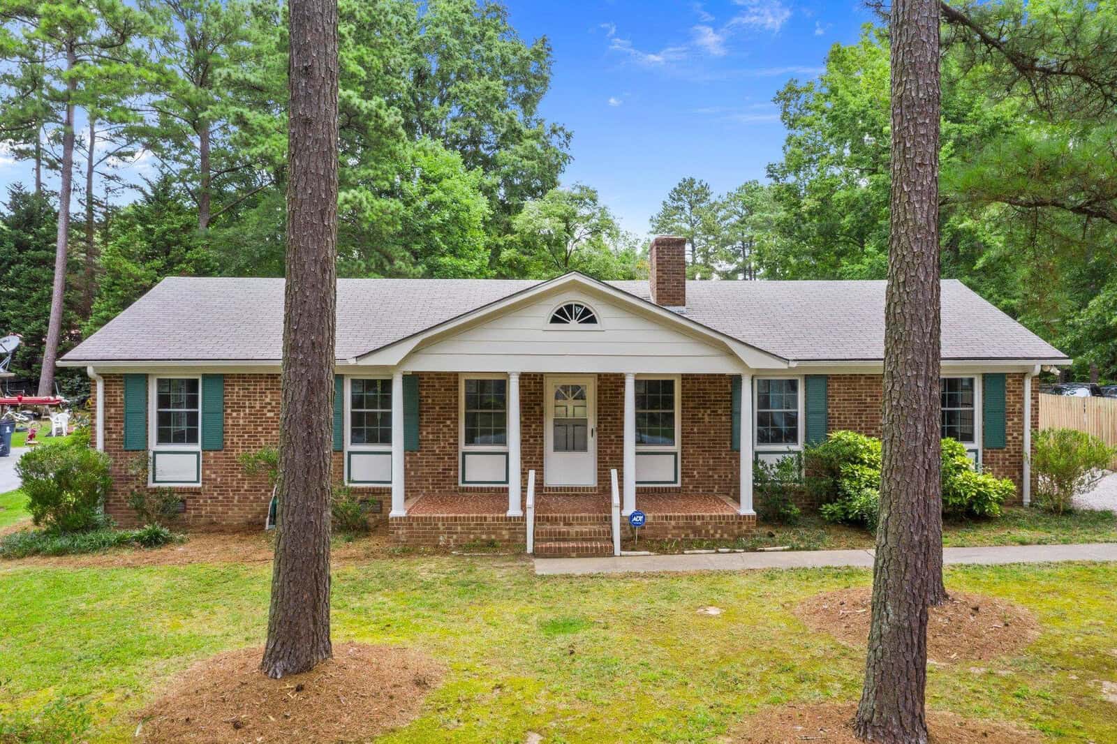 Single-story brick house with white front door and gable, surrounded by trees under a clear sky.