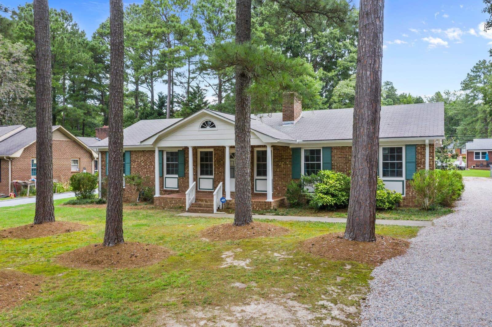 A single-story suburban home with a brick facade, white trim, and a shingled roof, surrounded by pine trees and a lawn with patches of bare earth. a gravel driveway leads up to the house.