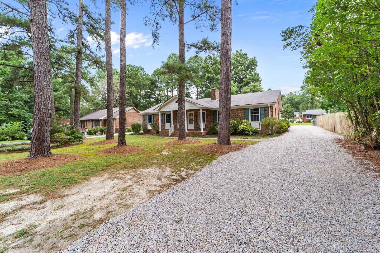 A single-story house with a brown brick exterior, nestled among tall pine trees, featuring a gravel driveway leading up to the double garage doors.