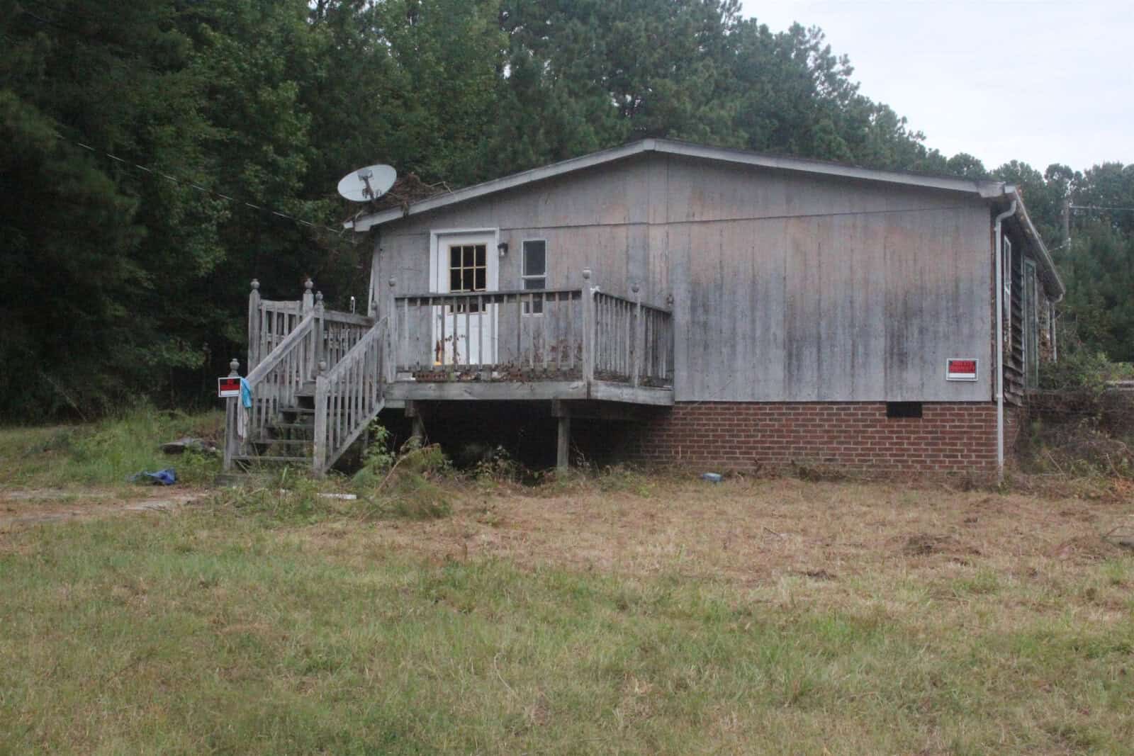 A weathered house with a wooden deck and brick foundation sits in a grassy area, with a satellite dish attached and various objects scattered on the ground.