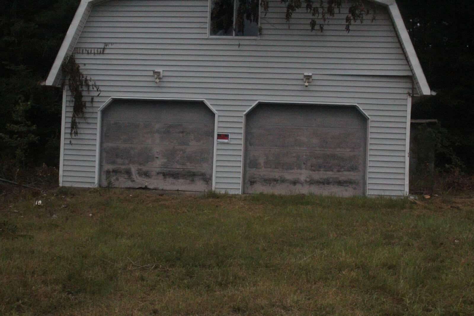 A white garage with two dirty doors, surrounded by a grassy area and some foliage in the background.