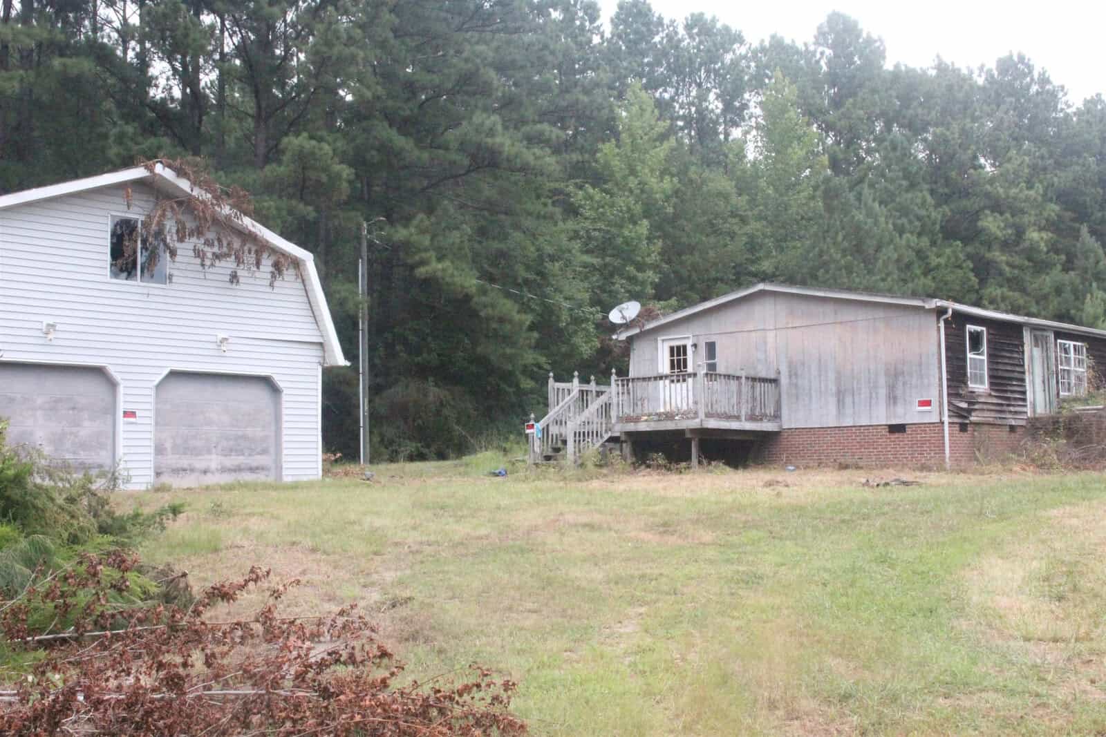 A weathered mobile home with an attached wooden deck alongside a white garage with overgrown foliage on the premises.