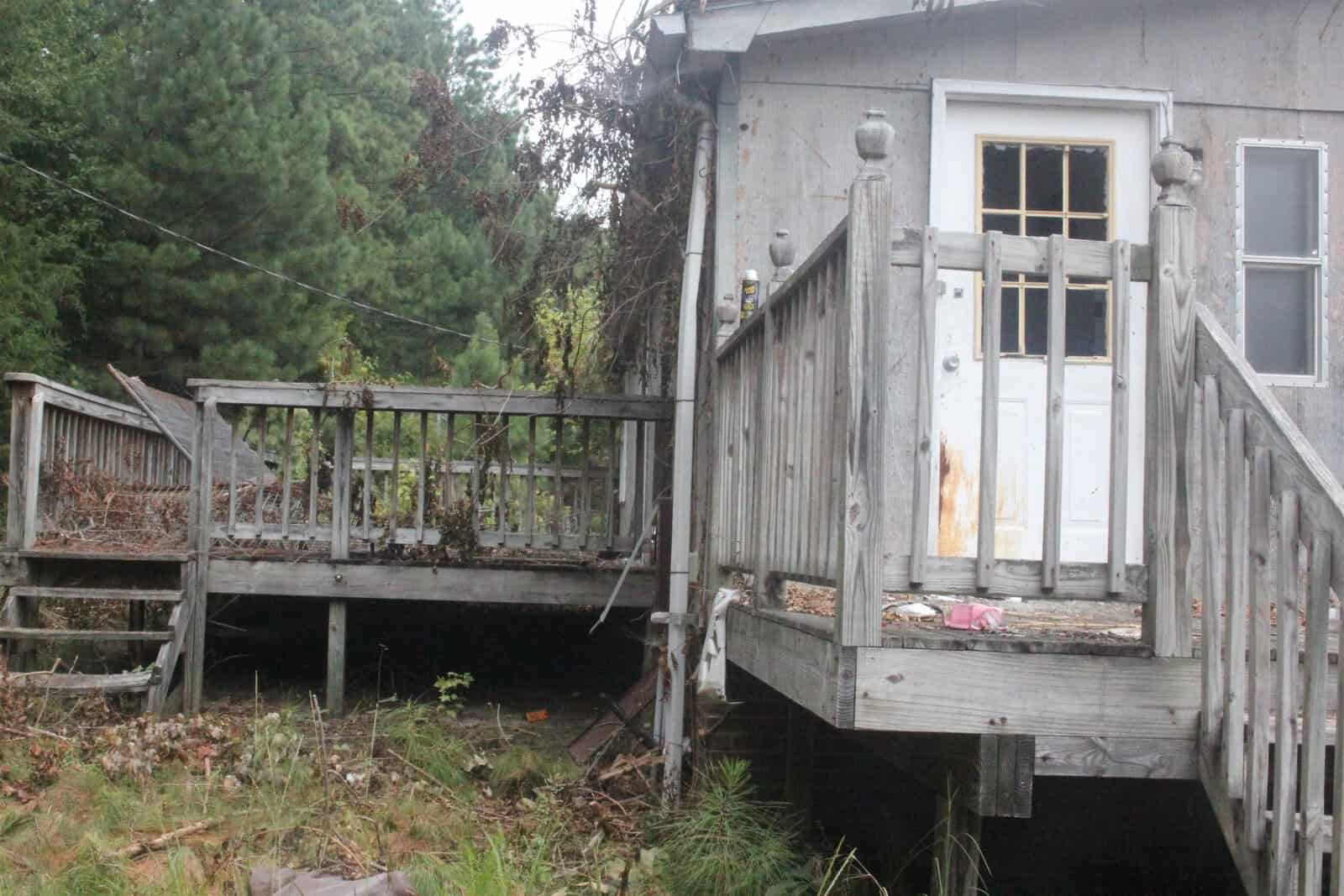A weathered wooden deck attached to a building with a white door, showing signs of disrepair and neglect, surrounded by overgrown vegetation.