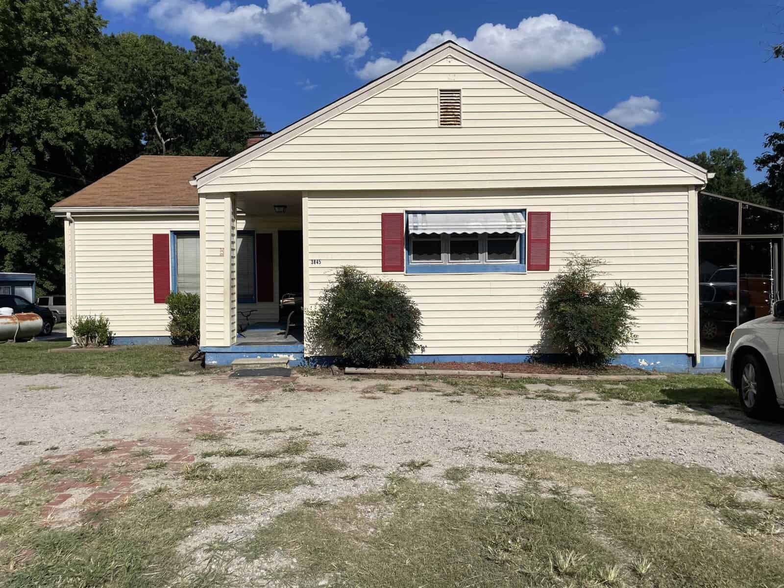 A single-story beige house with red shutters, a front porch, a driveway on the right side, and two small bushes flanking the front steps, under a clear blue sky.