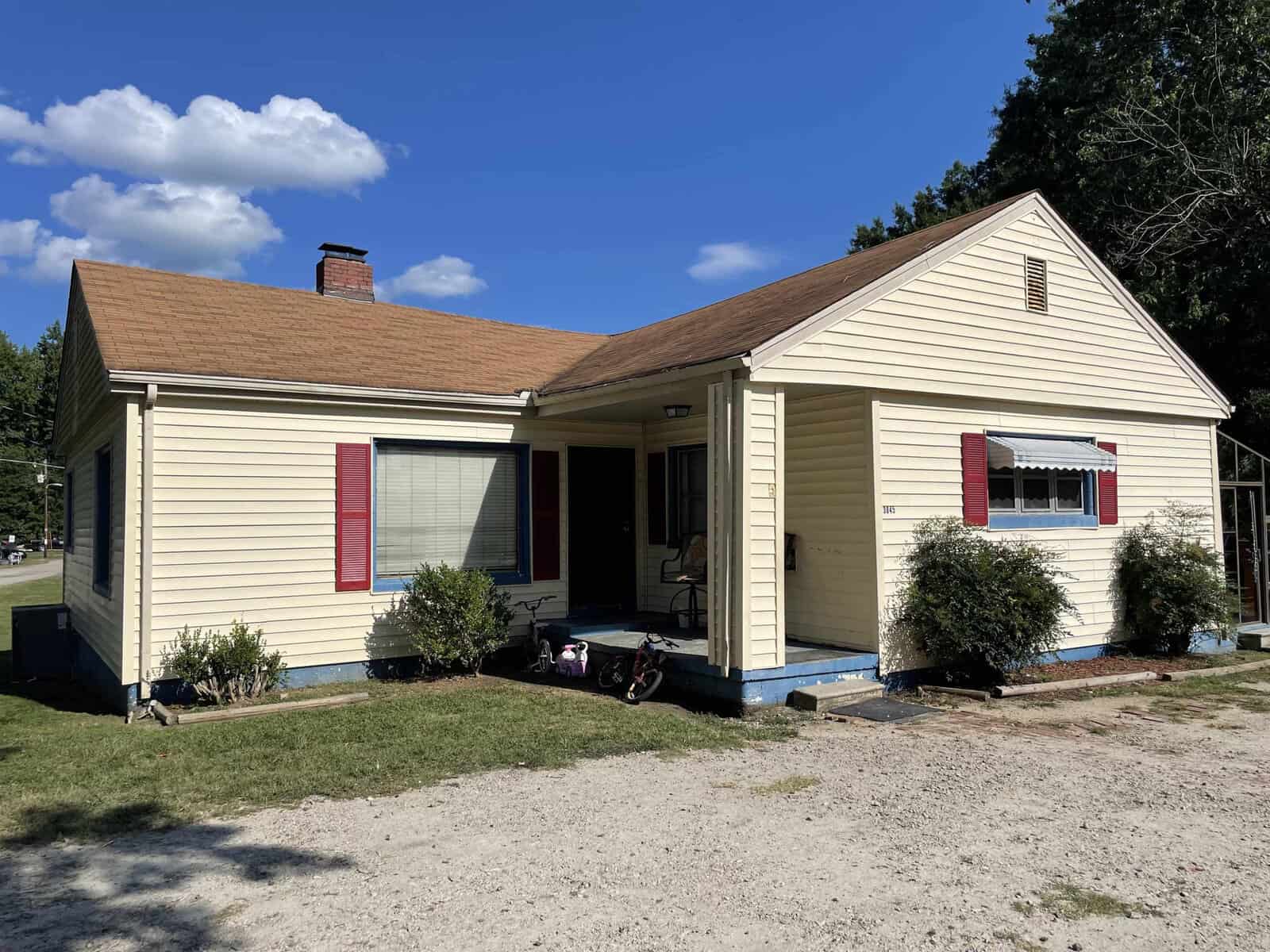 A single-story yellow house with red shutters, a front porch, and a lawn mower on the side.
