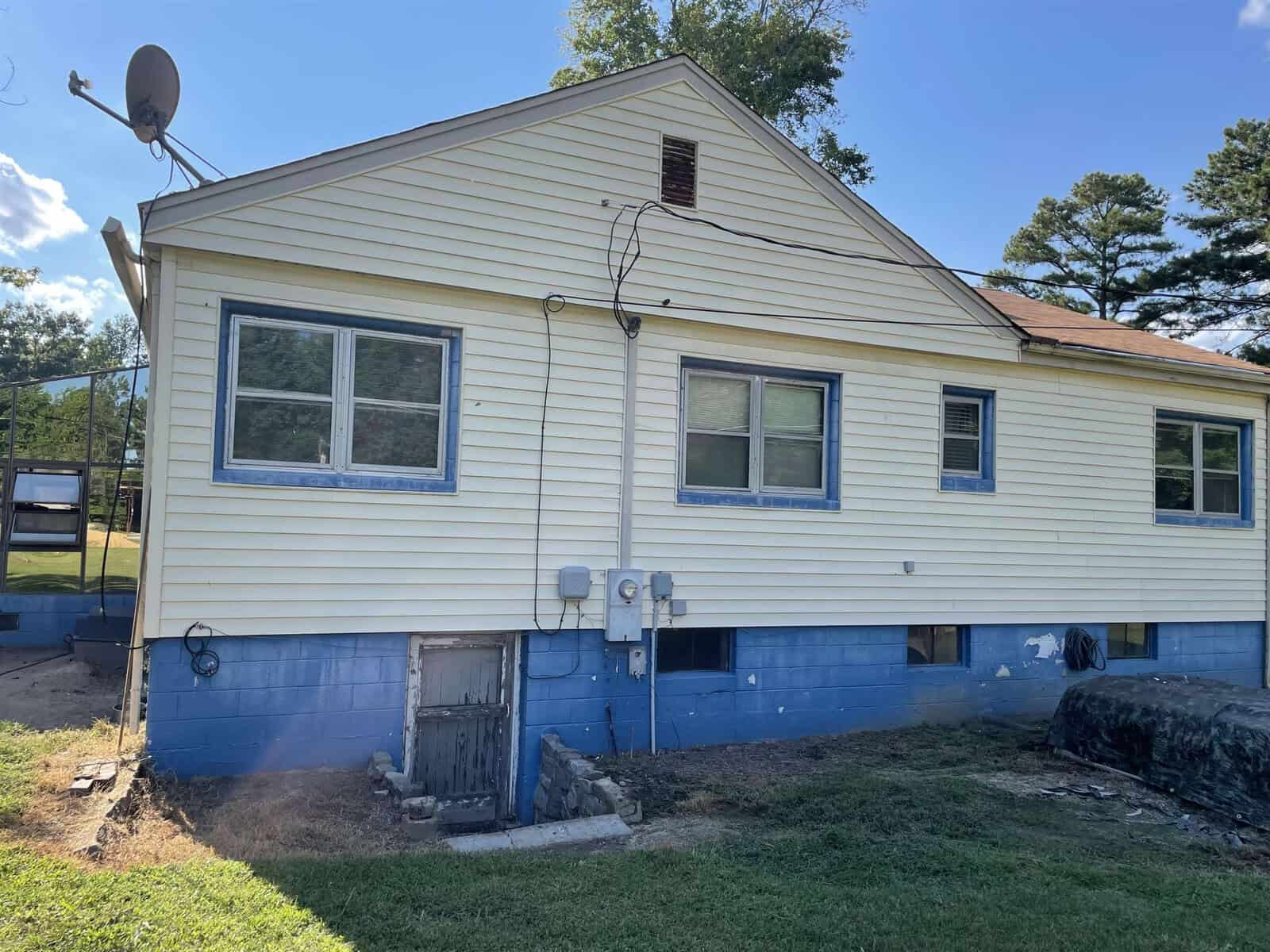 A single-story house with yellow siding and a blue foundation featuring a set of windows, an exterior utility box, and a small access door to the basement area, situated under a clear sky.