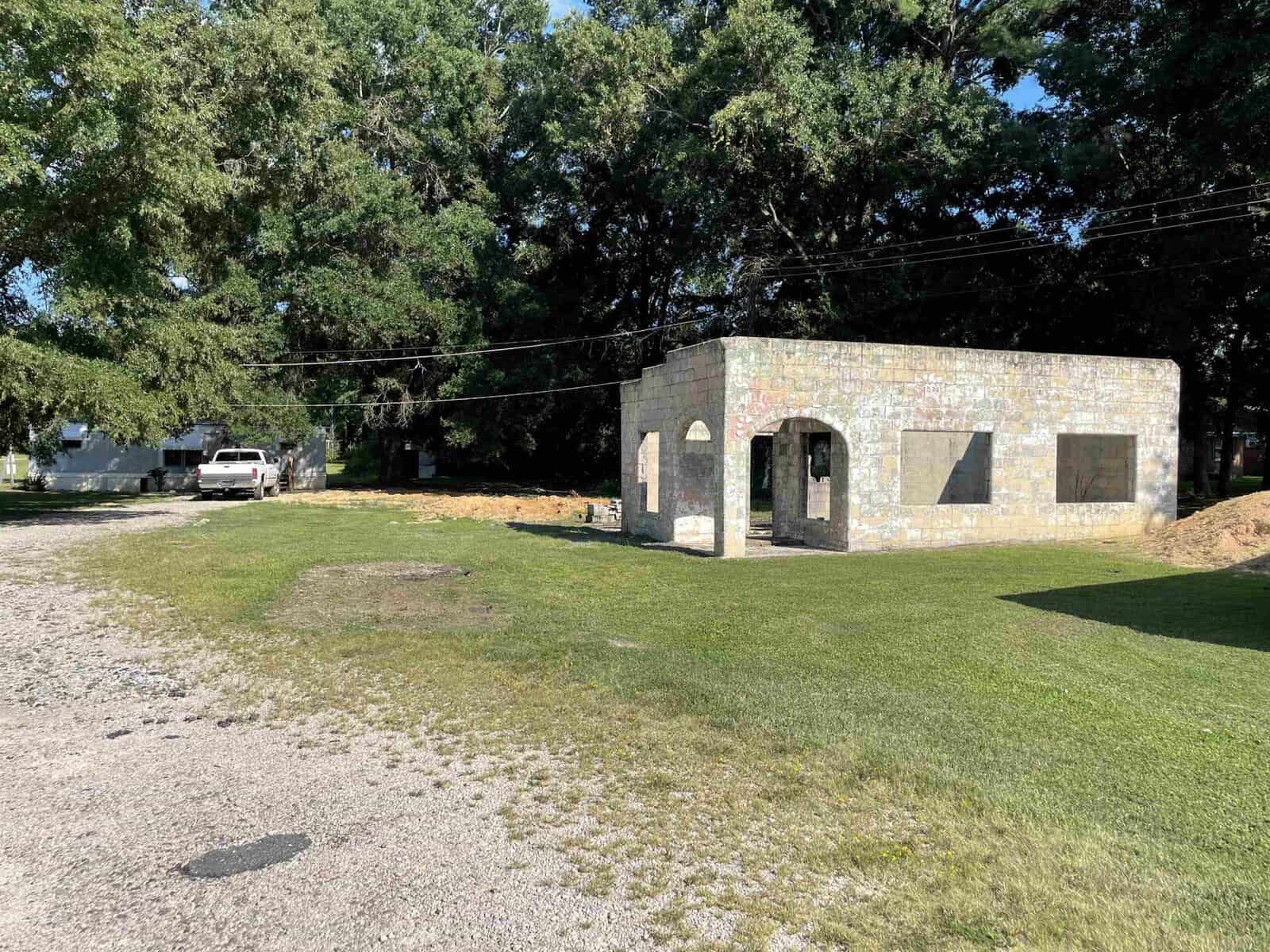 A small, single-story stone building with boarded-up windows sits under the shade of trees next to an open dirt area with a parked white vehicle.
