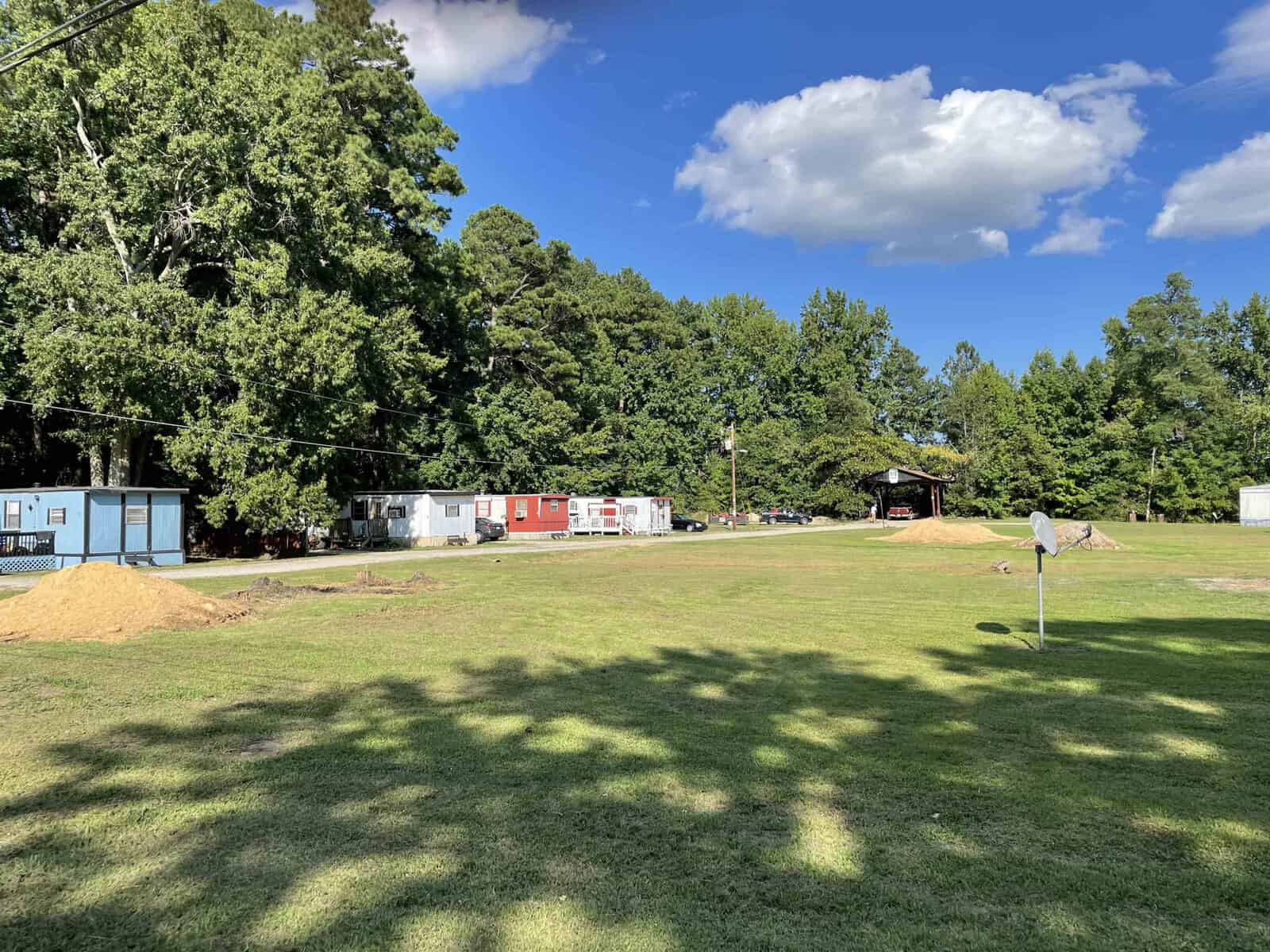 A sunny day at a park with scattered trees casting shadows on the grass, a basketball hoop in the foreground, and a row of small buildings in the background.