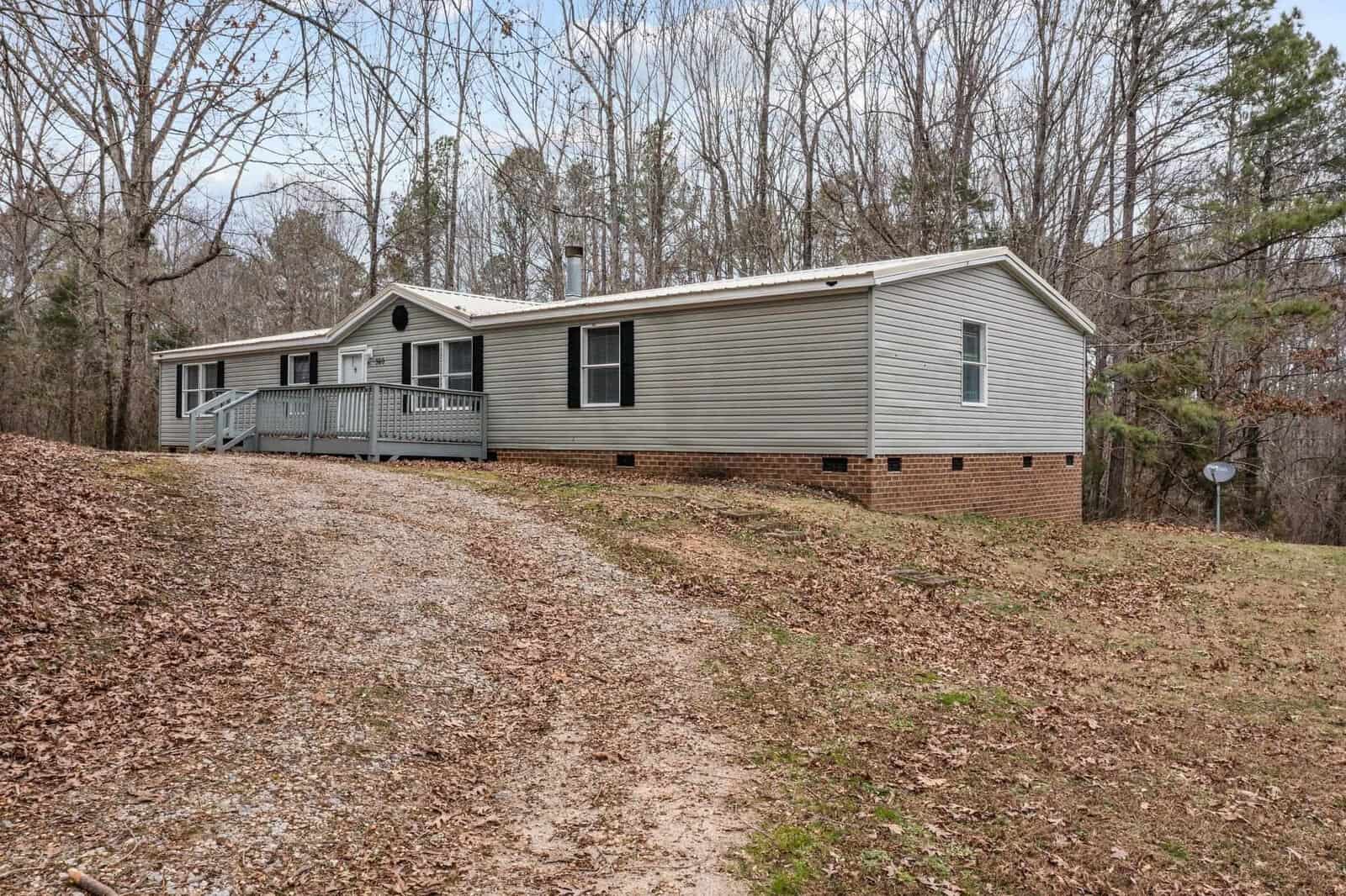 A single-story beige modular home with a small front porch and brick underpinning, situated on a sloped grassy terrain at 360 Spring Lane, Henderson NC. A gravel driveway leads to the