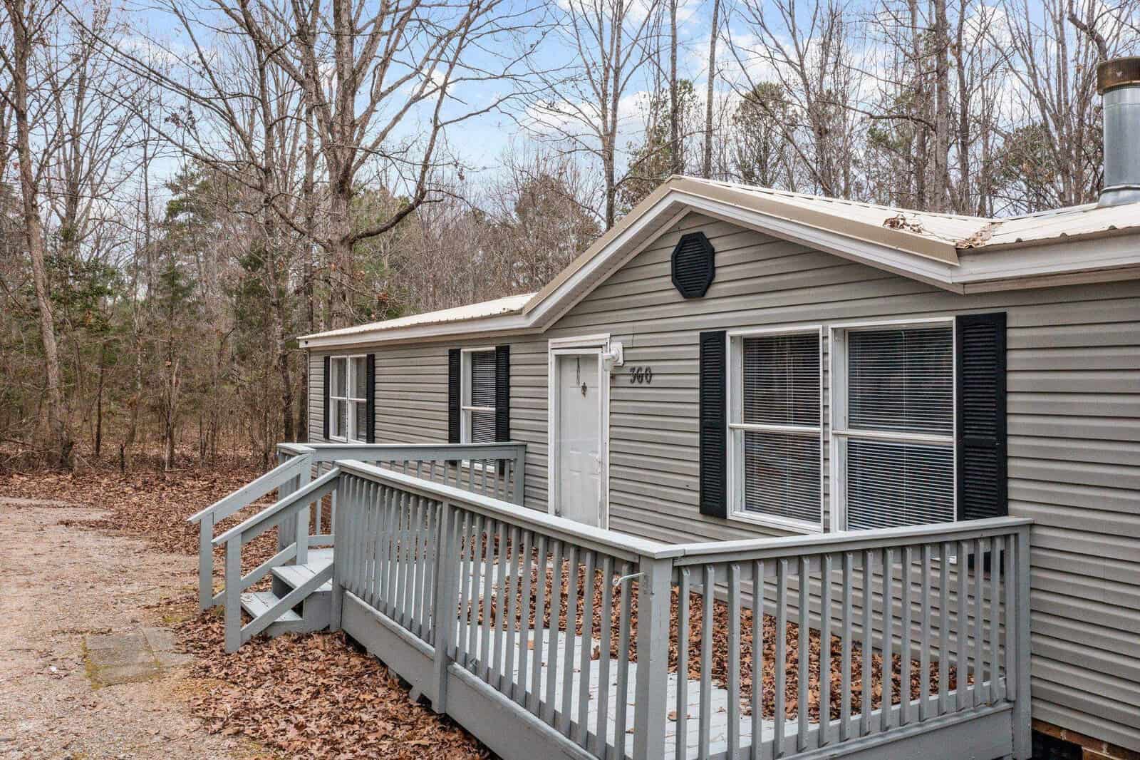 A single-story gray mobile home with a small wooden deck, surrounded by bare trees indicative of winter or early spring.