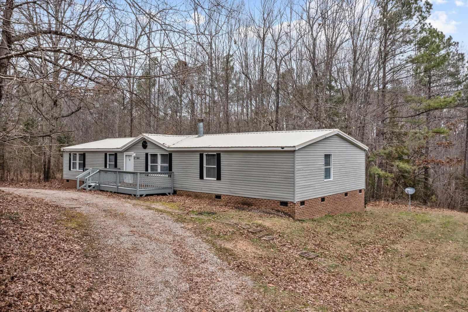 A single-story modular home with a gray exterior, featuring a small front porch and surrounded by a wooded area, with a gravel driveway leading up to the house.