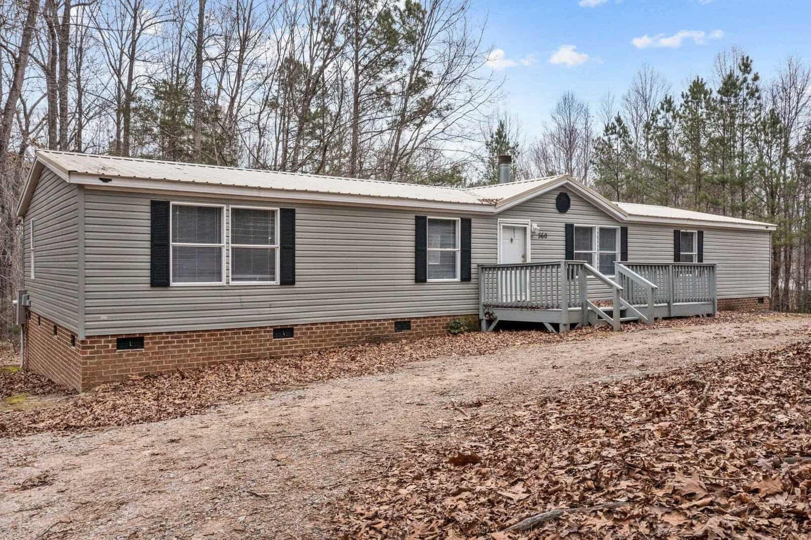 A single-story manufactured home with a gray exterior and brick underpinning, featuring a small front porch with steps, surrounded by bare trees and situated on a plot with scattered leaf litter.