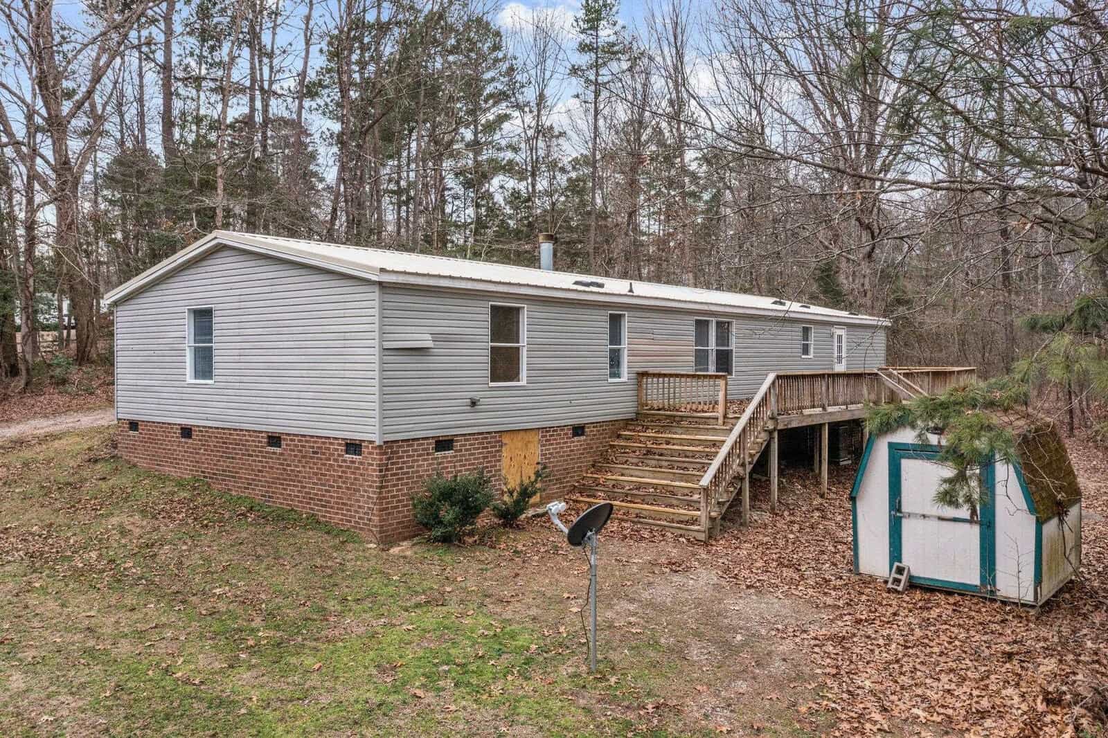 A single-story modular home with vinyl siding, featuring a small wooden deck and stairs, surrounded by bare trees and a modest lawn, with a small outdoor shed to the side.