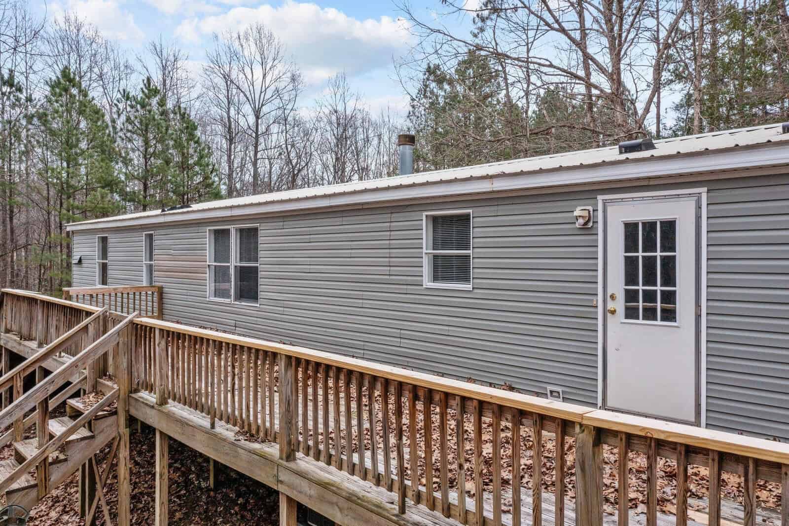 A single-story gray modular home with a white door, several windows, and an attached wooden deck surrounded by leafless trees.