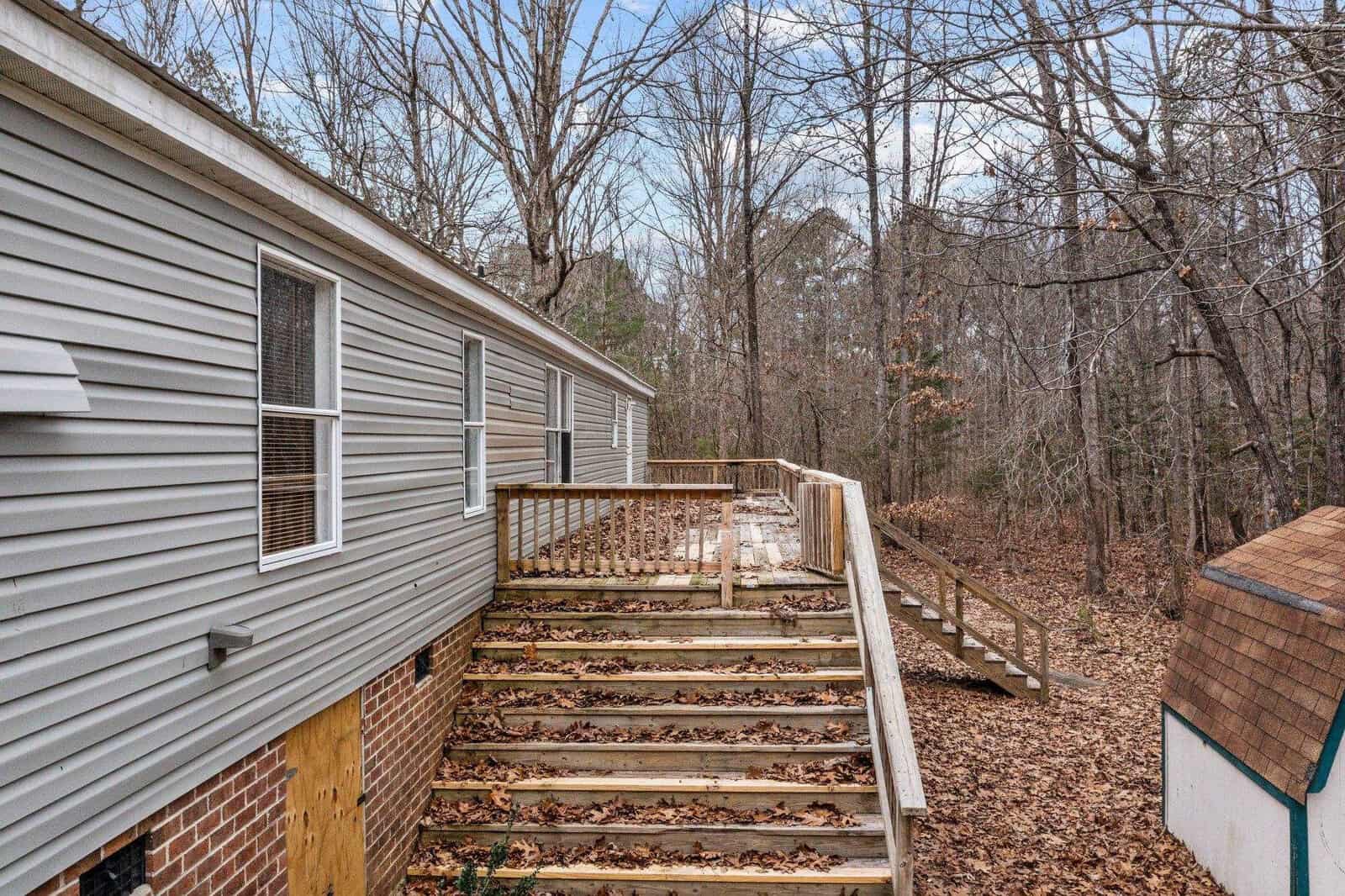 A side view of a gray mobile home with a wooden staircase and railing, surrounded by leafless trees and underbrush.