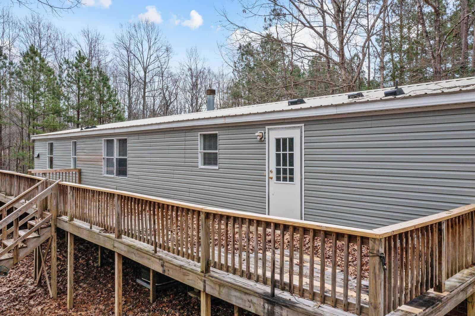 A single-story mobile home with gray siding, featuring a white door and a wooden deck, surrounded by leafless trees.
