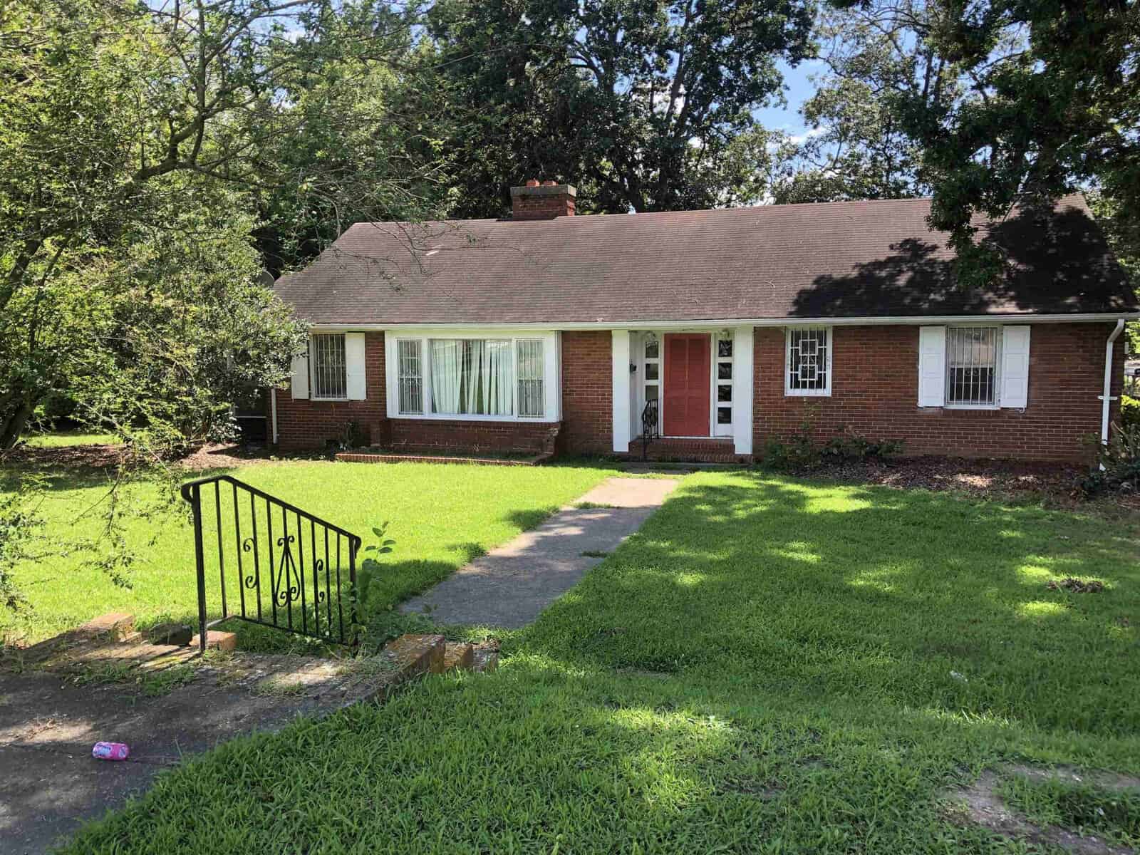 A single-story brick house on 1122 E Andrews Avenue, Henderson NC, with white-trimmed windows, a red front door, and a small concrete pathway leading to the entrance, surrounded by