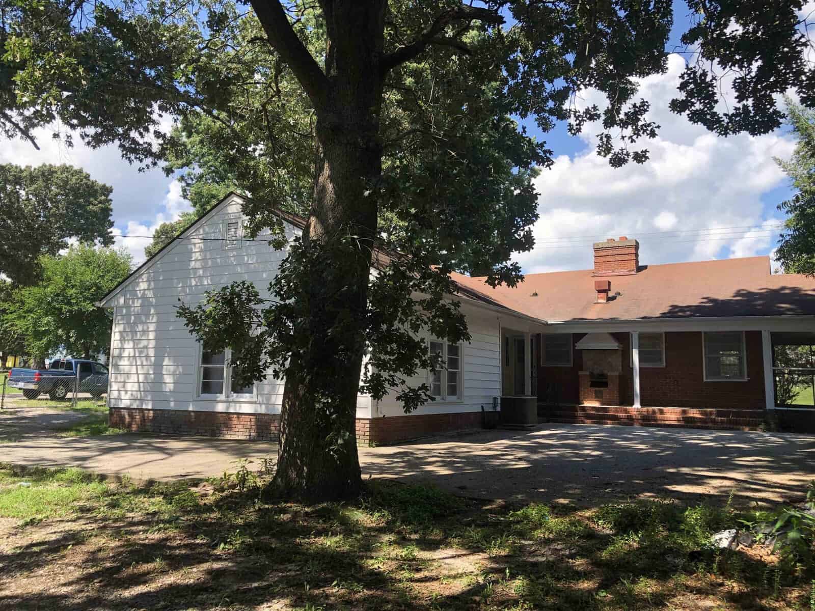 A detached single-story house with white siding and a brick facade under a clear sky, featuring a prominent tree in the front yard casting shadows on the driveway.