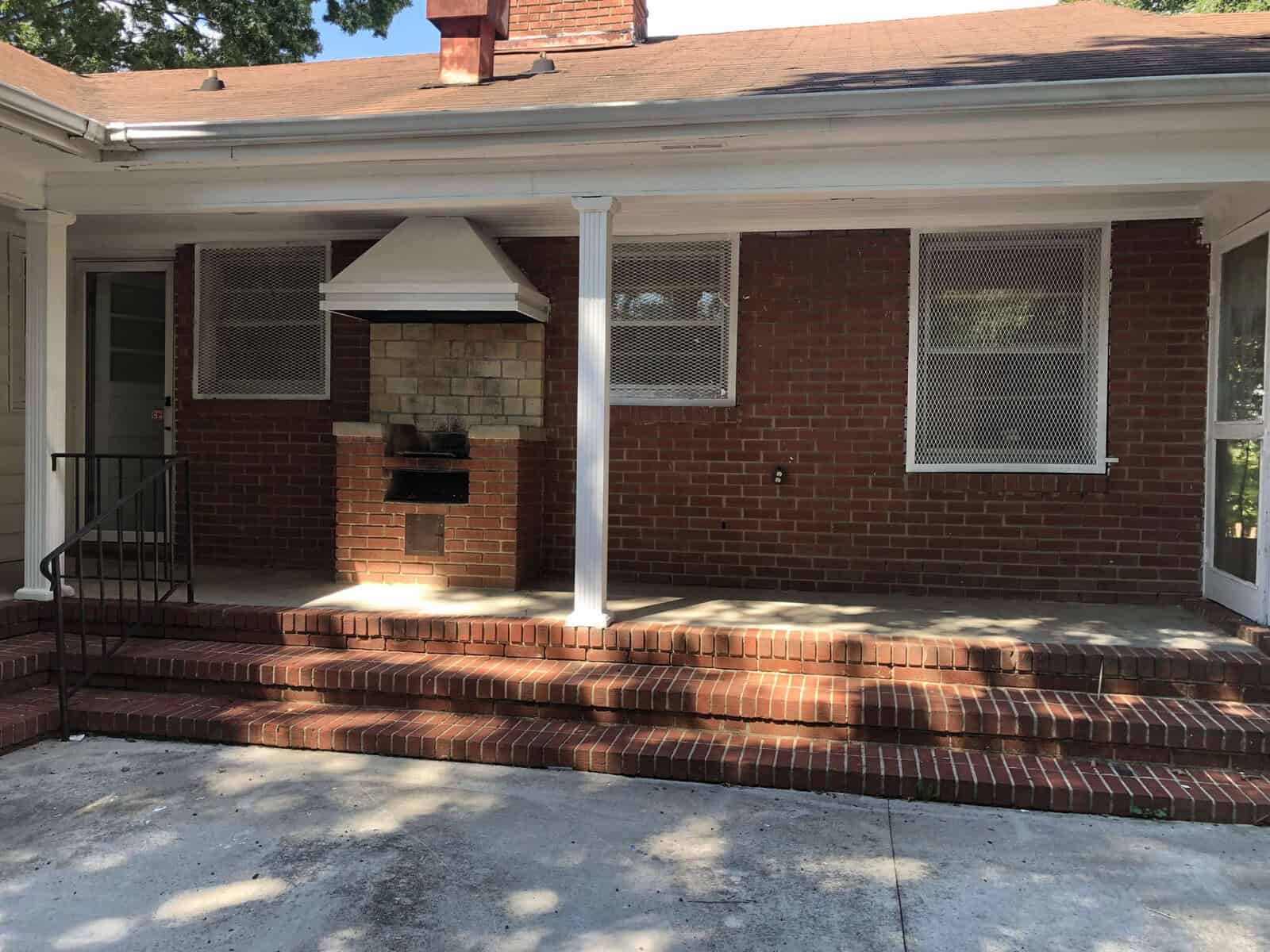 A traditional brick house featuring a small front porch with a built-in barbecue grill, white columns, and window screens, bathed in sunlight.