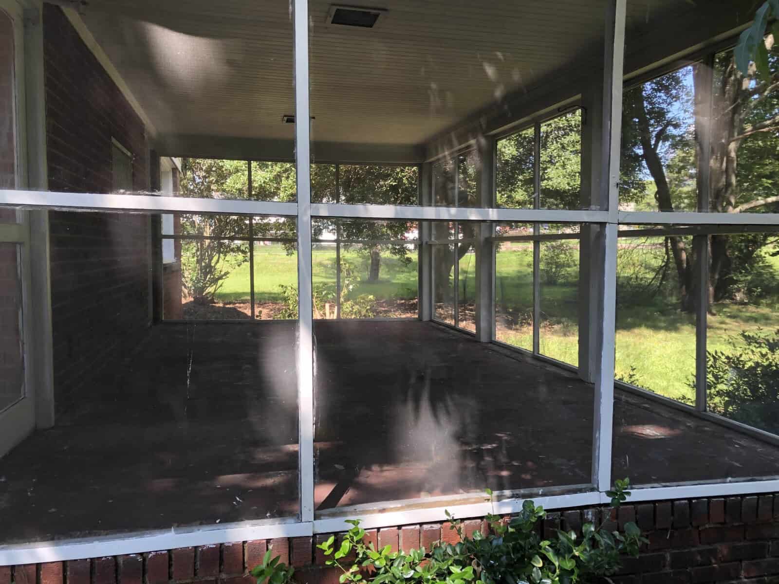 A screened porch with a concrete floor and a view of a green lawn with trees, attached to a brick house.