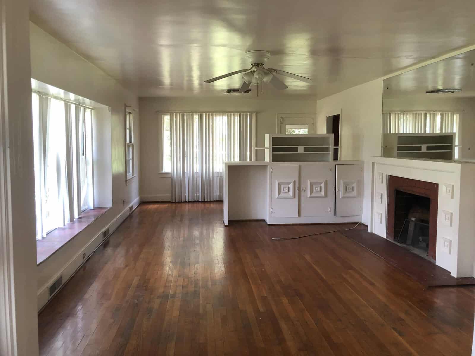 Empty living room with hardwood floors, a white fireplace, and large windows allowing natural light in.