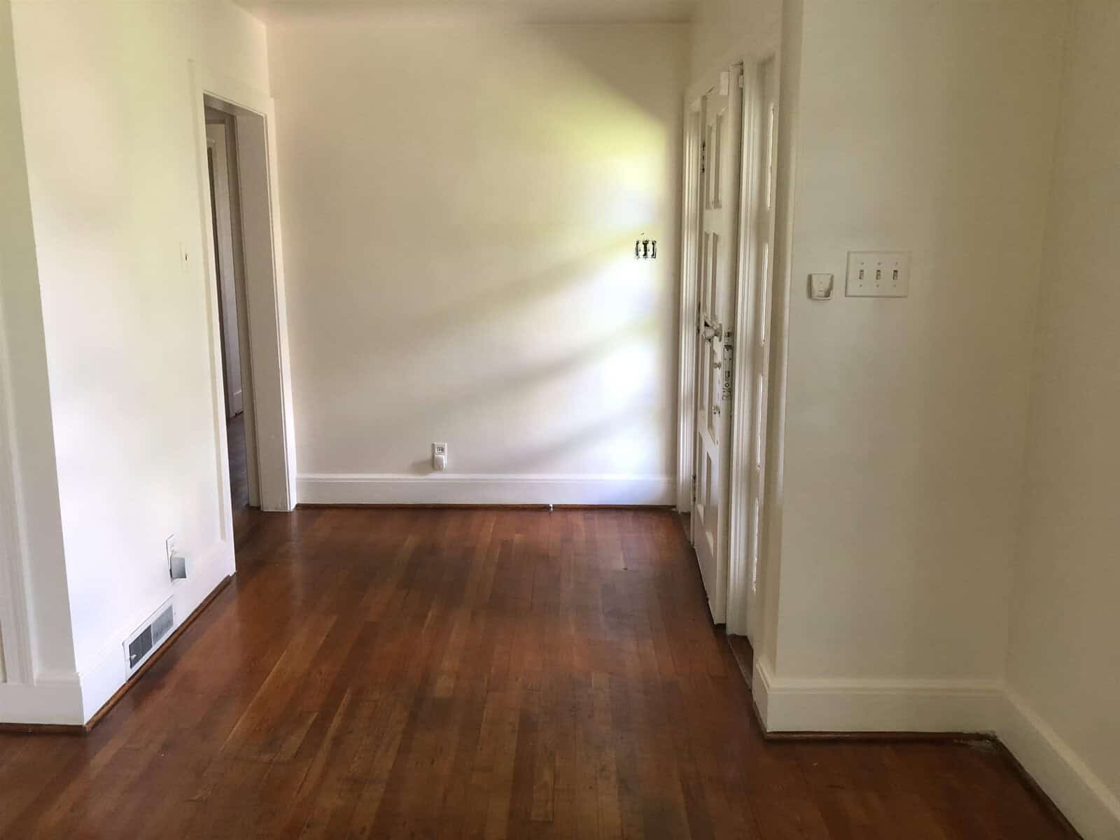 An empty room with hardwood flooring, white walls, and a closed white door, illuminated by natural light coming in from a window out of view.