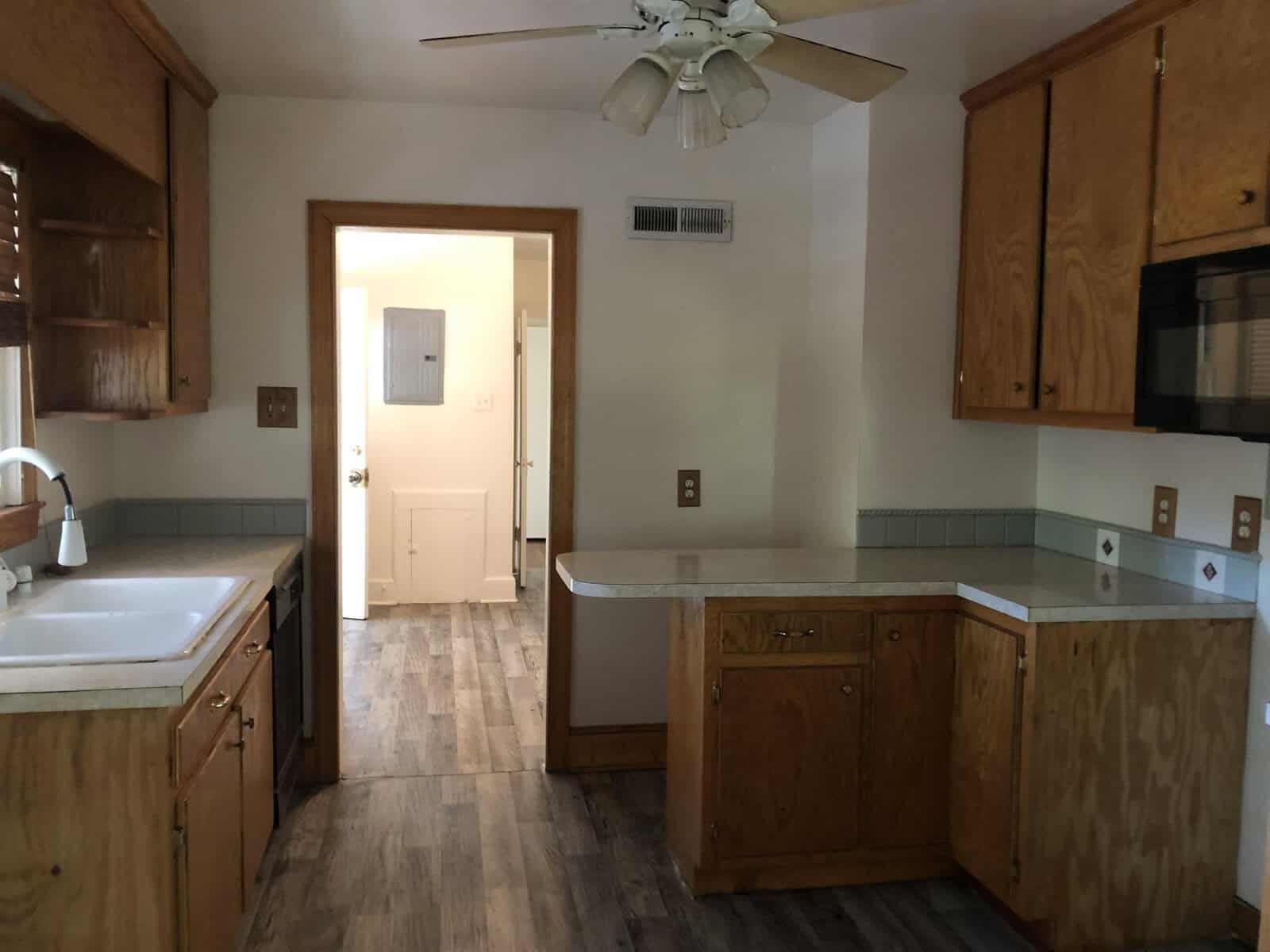 A spacious kitchen with wood cabinetry, laminate countertops, and vinyl flooring, featuring a sink with no faucet installed and unoccupied electrical outlets, highlighted by natural light and a ceiling fan overhead.
