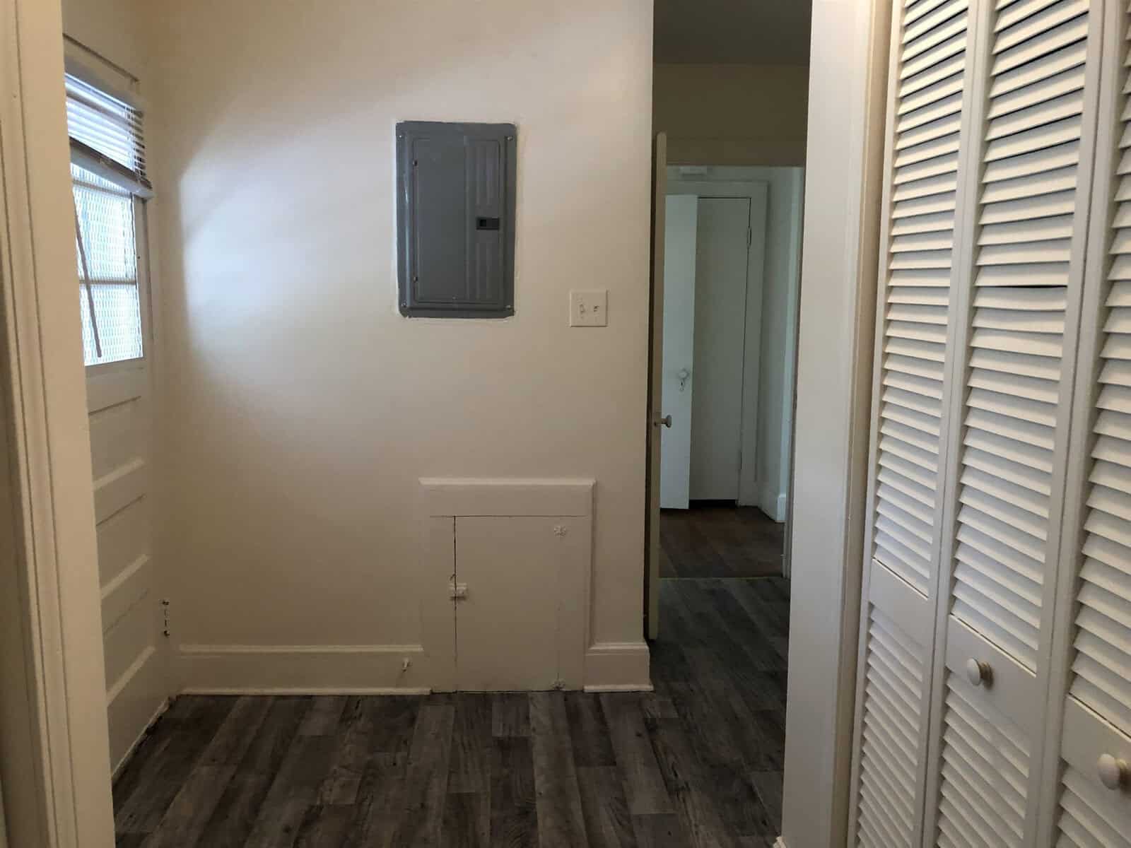 An empty room corner with beige walls, featuring a small closed door, presumably accessing under-stair storage, an electrical panel, and two white louvered doors likely leading to a closet, with another room visible through an open doorway. the floor is covered with wood-patterned vinyl or laminate flooring.