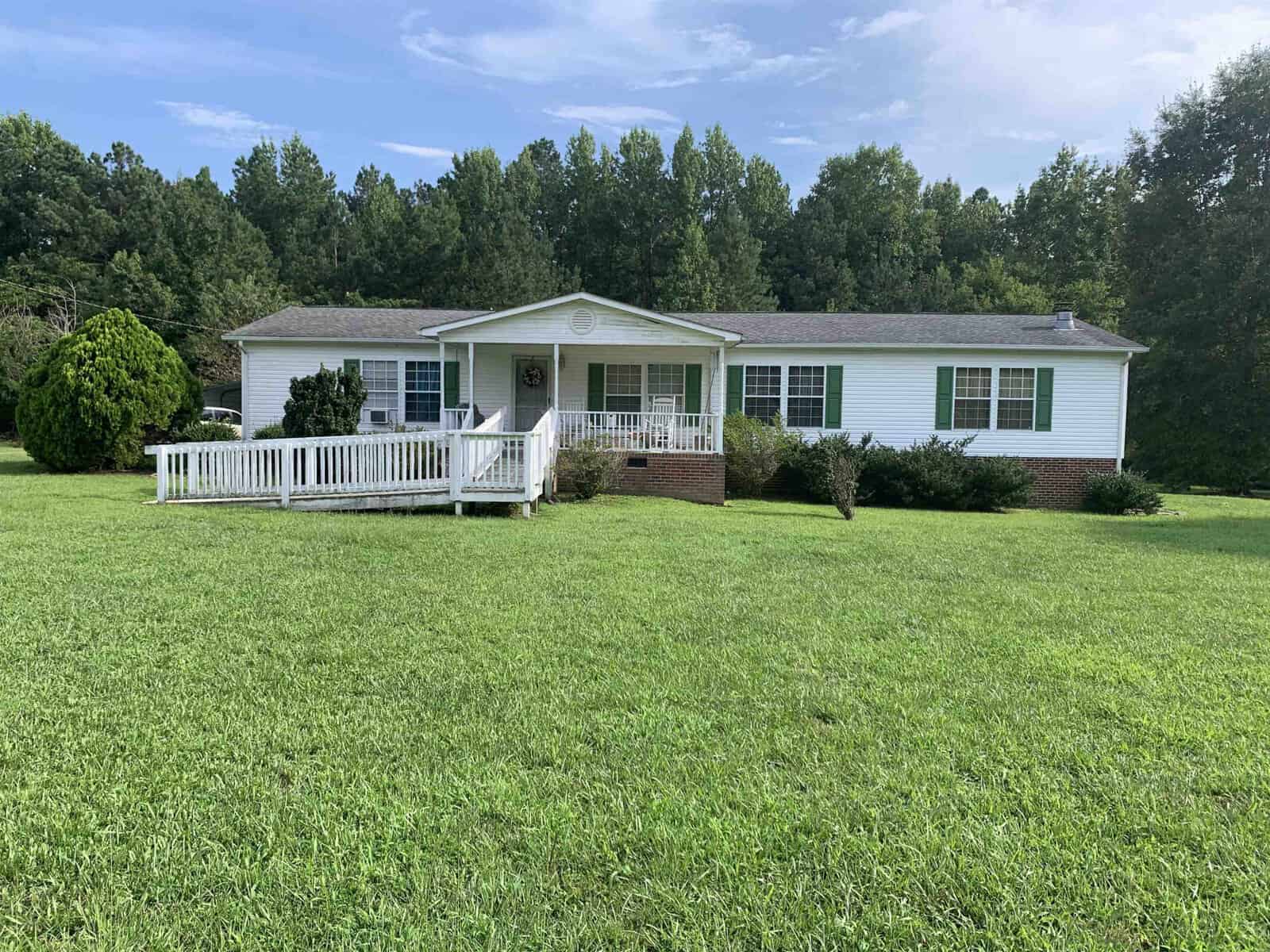 A white, single-story house on 983 Bullocksville Park Road, Manson, features a wheelchair-accessible ramp and lush green lawn. The foreground showcases a spacious grassy area, while the backdrop is adorned with tall trees under a partly cloudy sky.