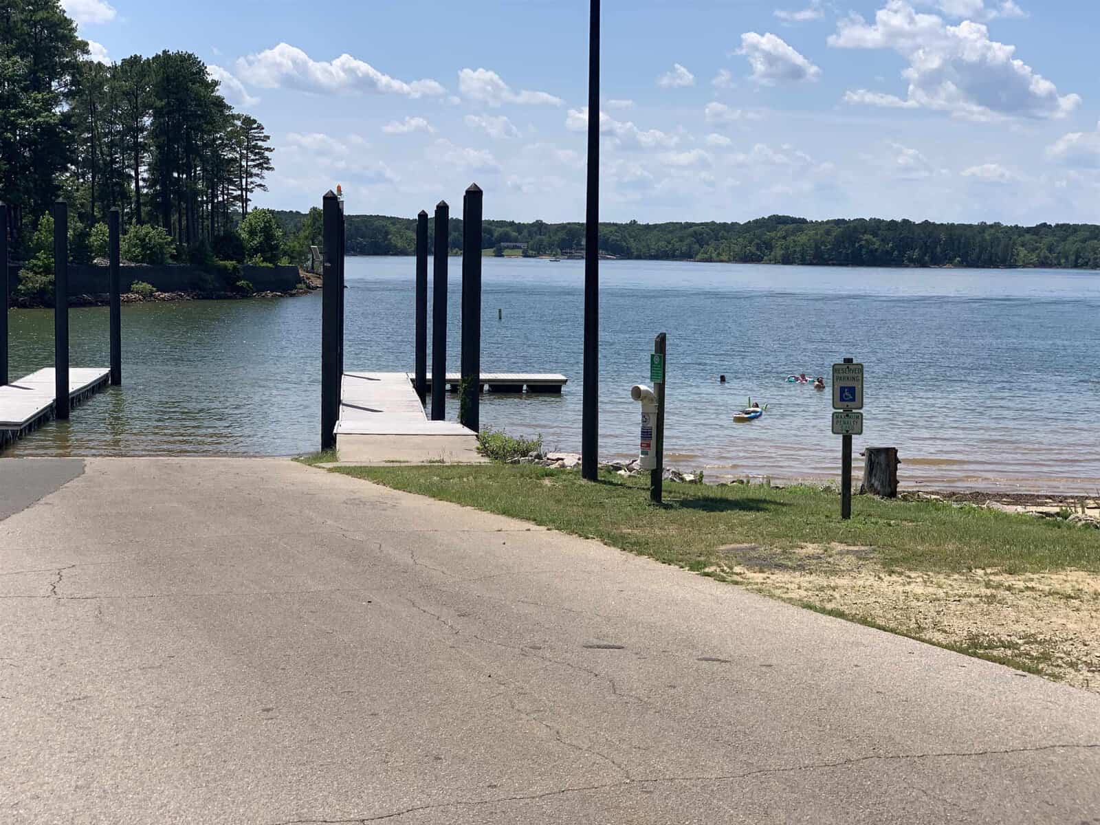 A boat launch ramp leads to a lake with a dock extending into the water. Several people swim nearby under a partly cloudy sky. Trees line the distant shore, and a sign with accessibility symbols is visible by the path.