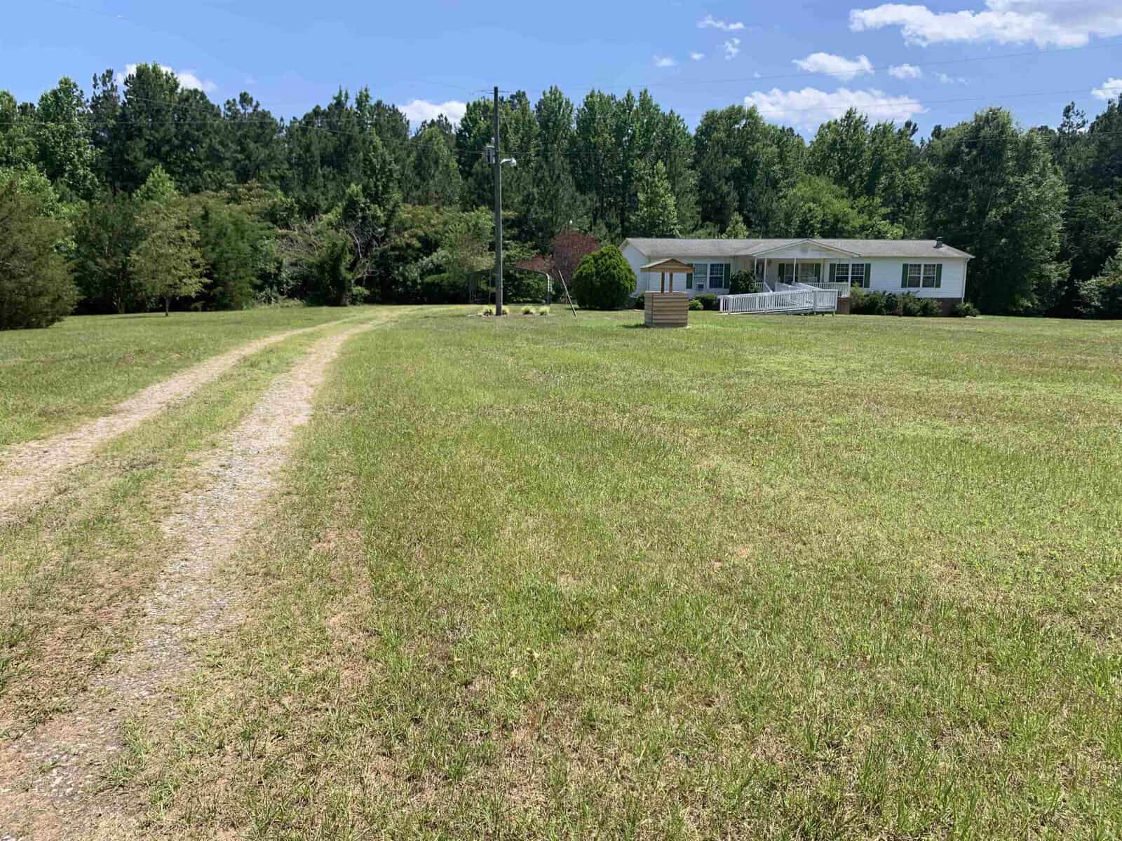 A rural landscape featuring a gravel path leading to a small white house with a wooden porch. The house is surrounded by a large green lawn and trees under a bright blue sky with scattered clouds.