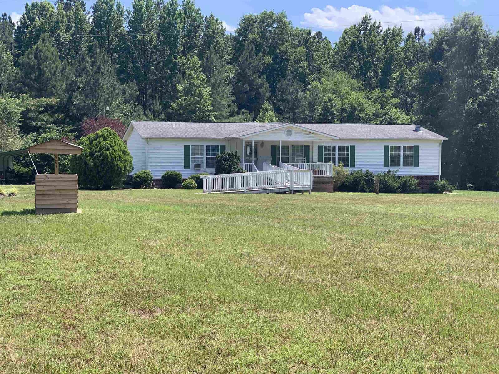 A white, single-story house with a front porch and ramp is surrounded by a large, grassy yard. Trees line the background under a clear blue sky. A wooden well structure is to the side.