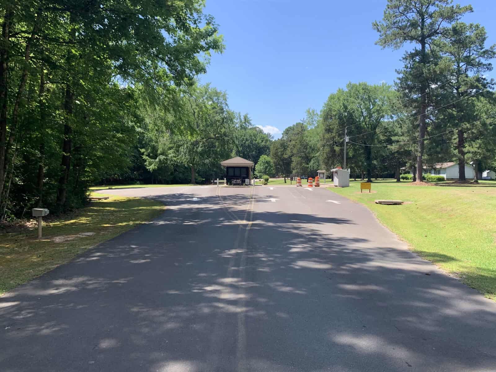 A paved road leads to a small wooden building surrounded by trees under a clear blue sky. To the right, there's an open grassy area and a couple of utility poles. Orange cones partially block the road near the building.
