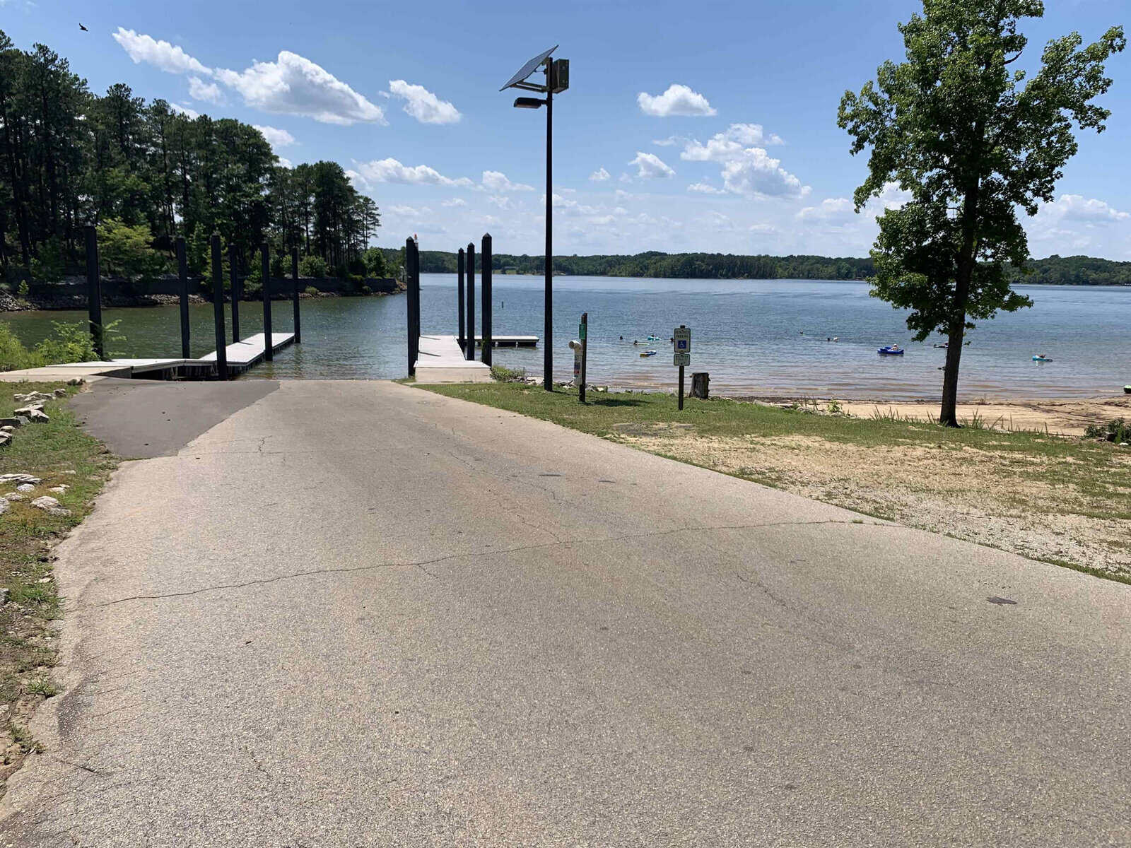 A paved boat ramp leads into a large lake under a clear blue sky with scattered clouds. The area is surrounded by trees, and there are a few boats visible on the water. A solar-powered streetlight stands beside the ramp.