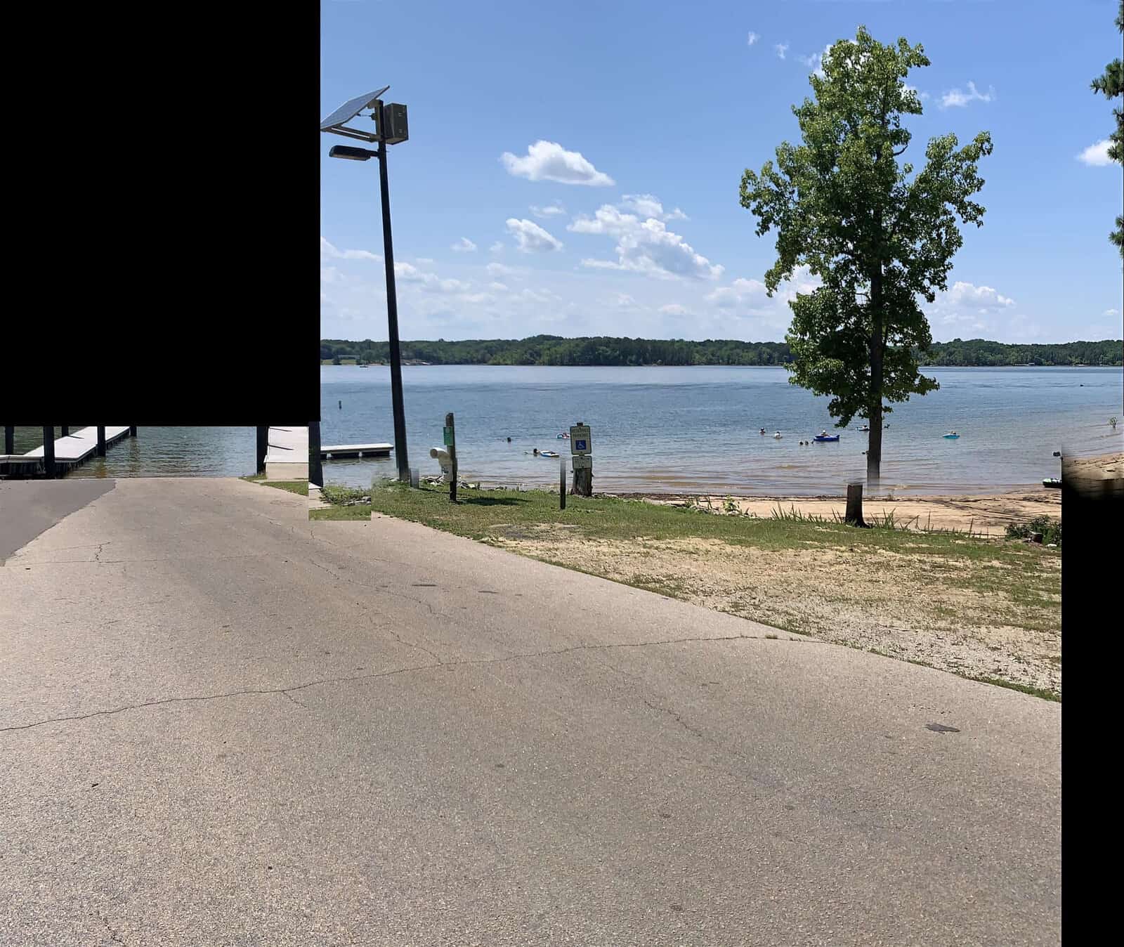 A serene lake scene with a paved road leading to the water, where people are swimming and kayaking. Trees border the lake, and the sky is scattered with fluffy clouds on a sunny day.