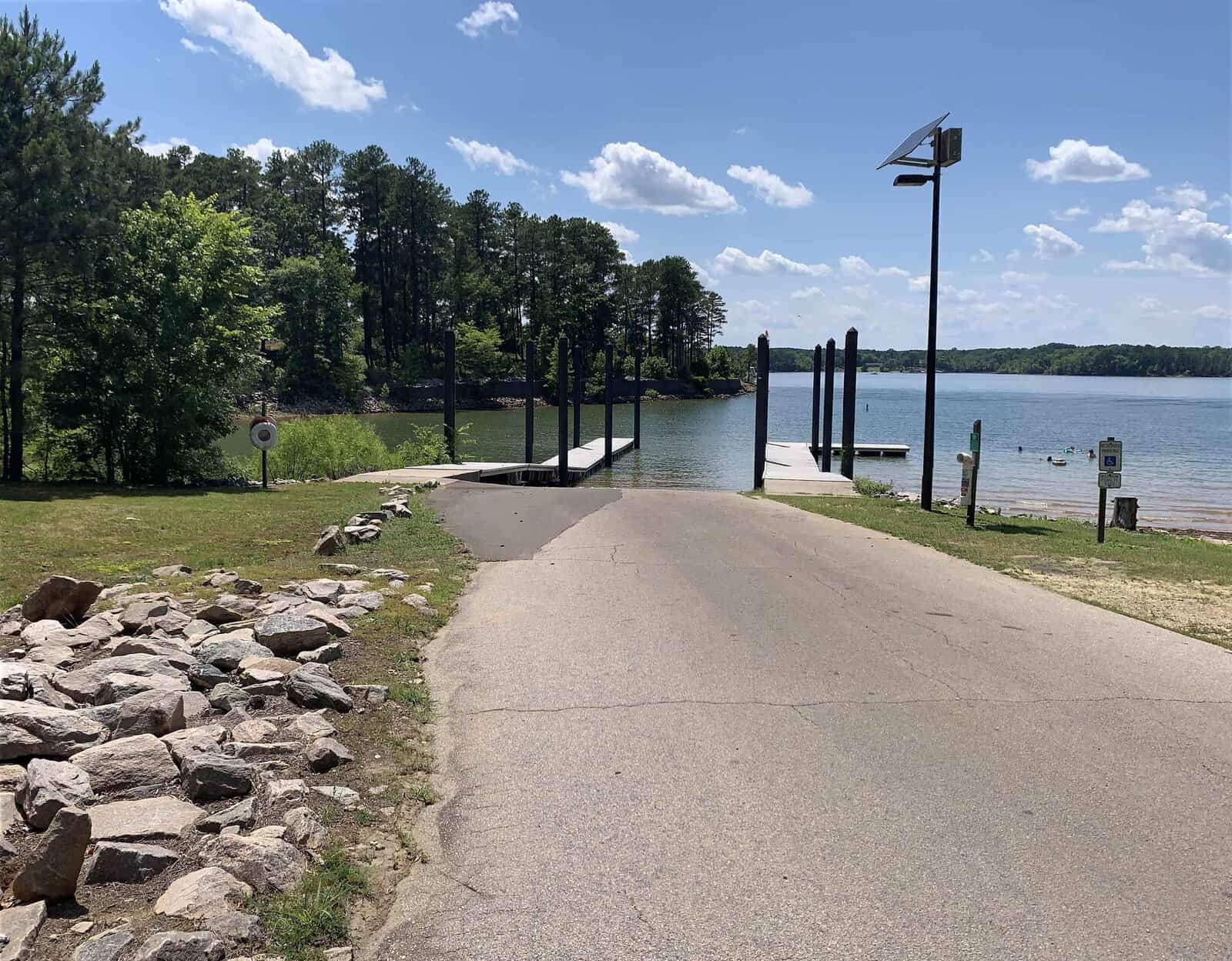 A sunny lakeside scene with a paved boat ramp leading into the water. Pine trees line the background, and several people are swimming near the shore. A solar-powered light pole stands nearby, and a rocky area is visible to the left.