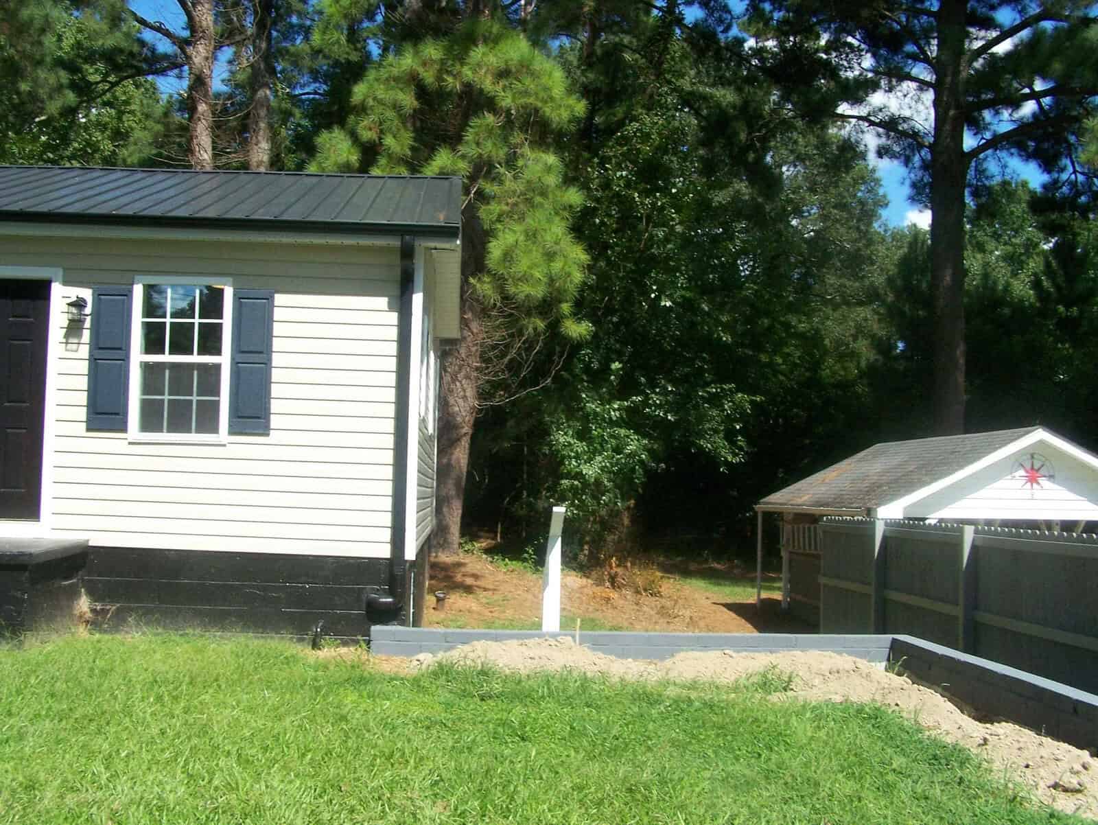 A suburban backyard featuring a gray-sided house with black shutters, a wood-fenced area on the right, and a small garden shed with a star decoration in the background amidst green trees.