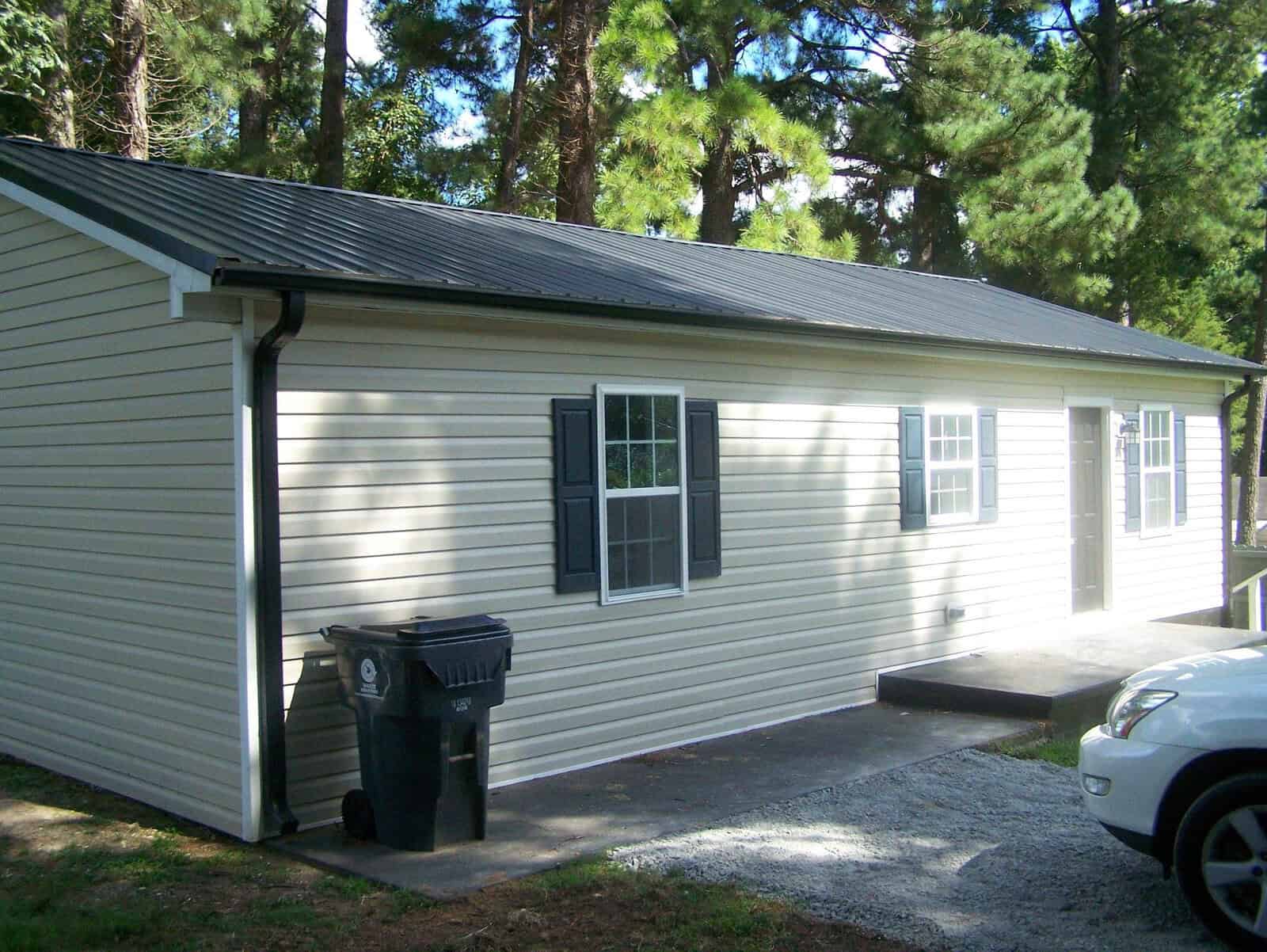 A single-story building with white sidings and a gabled roof, flanked by tall pine trees, with a black wheeled trash bin on the left and a parked car on the right.