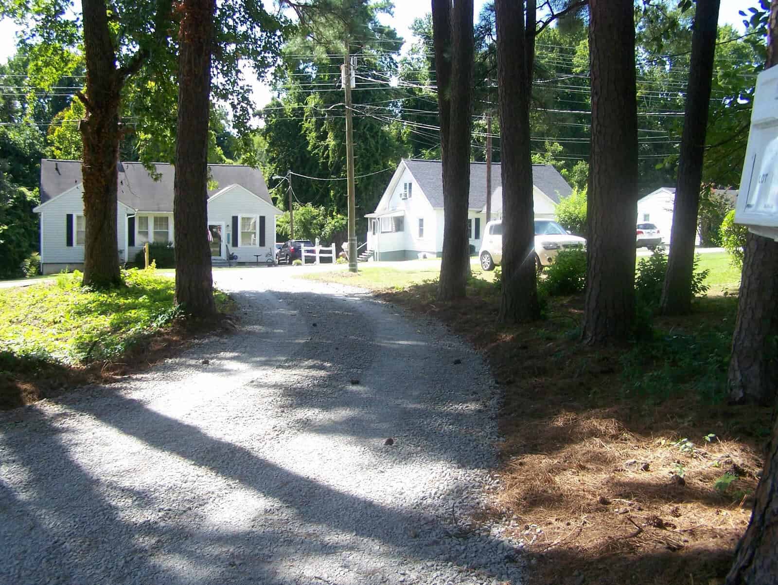 A residential street lined with trees and houses on a sunny day, with driveways leading to the homes and a mailbox in the foreground.