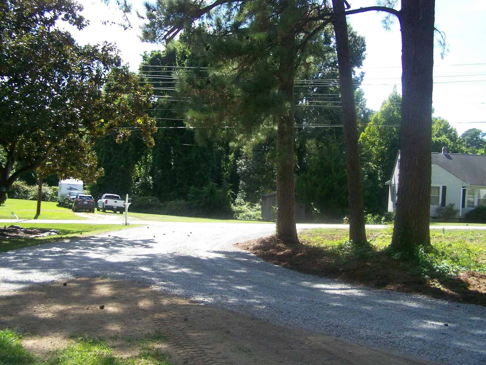 A sunny day on a residential street with trees and a gravel driveway; a white house and parked vehicles are visible in the background.