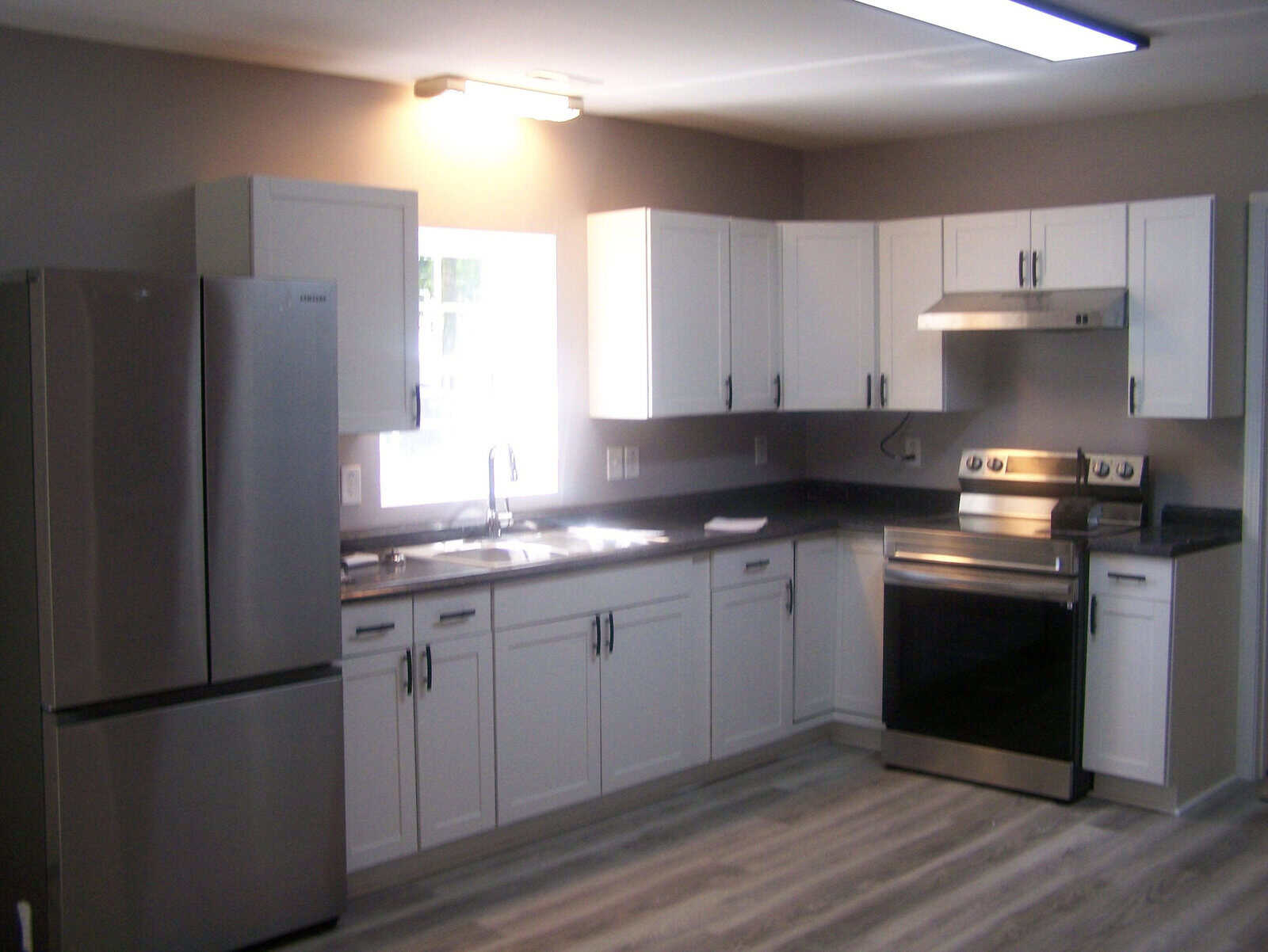 A modern kitchen with stainless steel appliances, white shaker cabinets, and dark countertops, featuring a refrigerator, dishwasher, sink, and an electric range with an overhead exhaust hood.