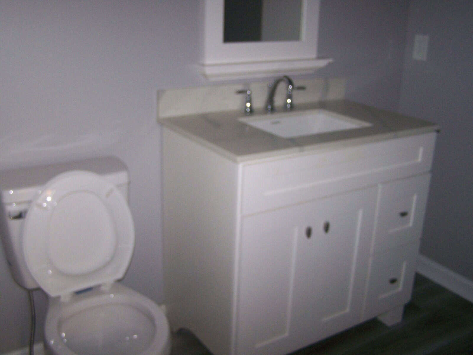 A bathroom interior with a white toilet to the left and a white vanity cabinet with a sink and faucet to the right, against a grey wall.