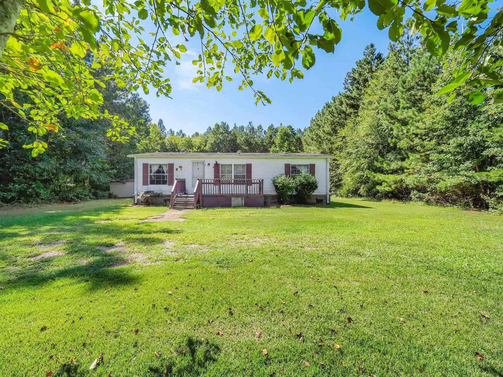 A single-story white house with brown shutters sits at 1207 Barker Road, Oxford. It features a wooden porch and is surrounded by lush trees under a clear blue sky, all set on a large green lawn.