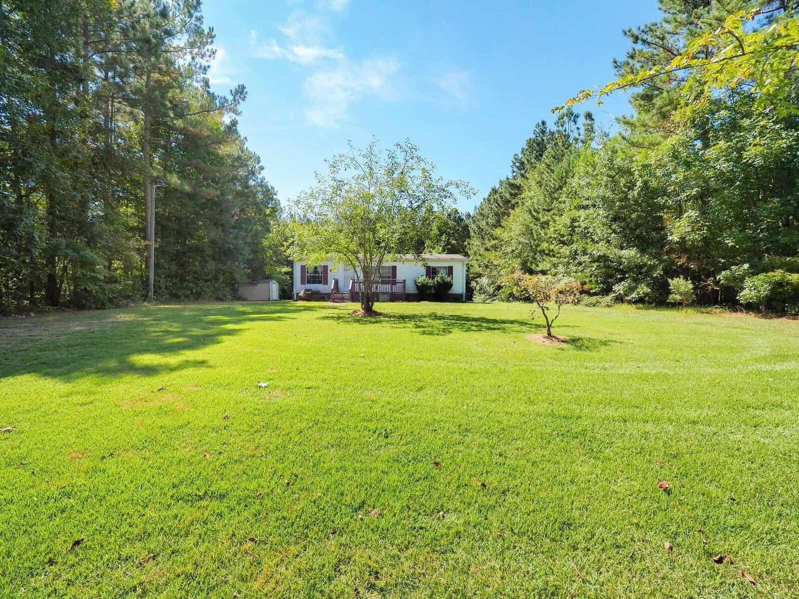 A white house with a porch is situated in the center of a lush green lawn, surrounded by tall trees under a clear blue sky. A small tree stands prominently in the foreground.