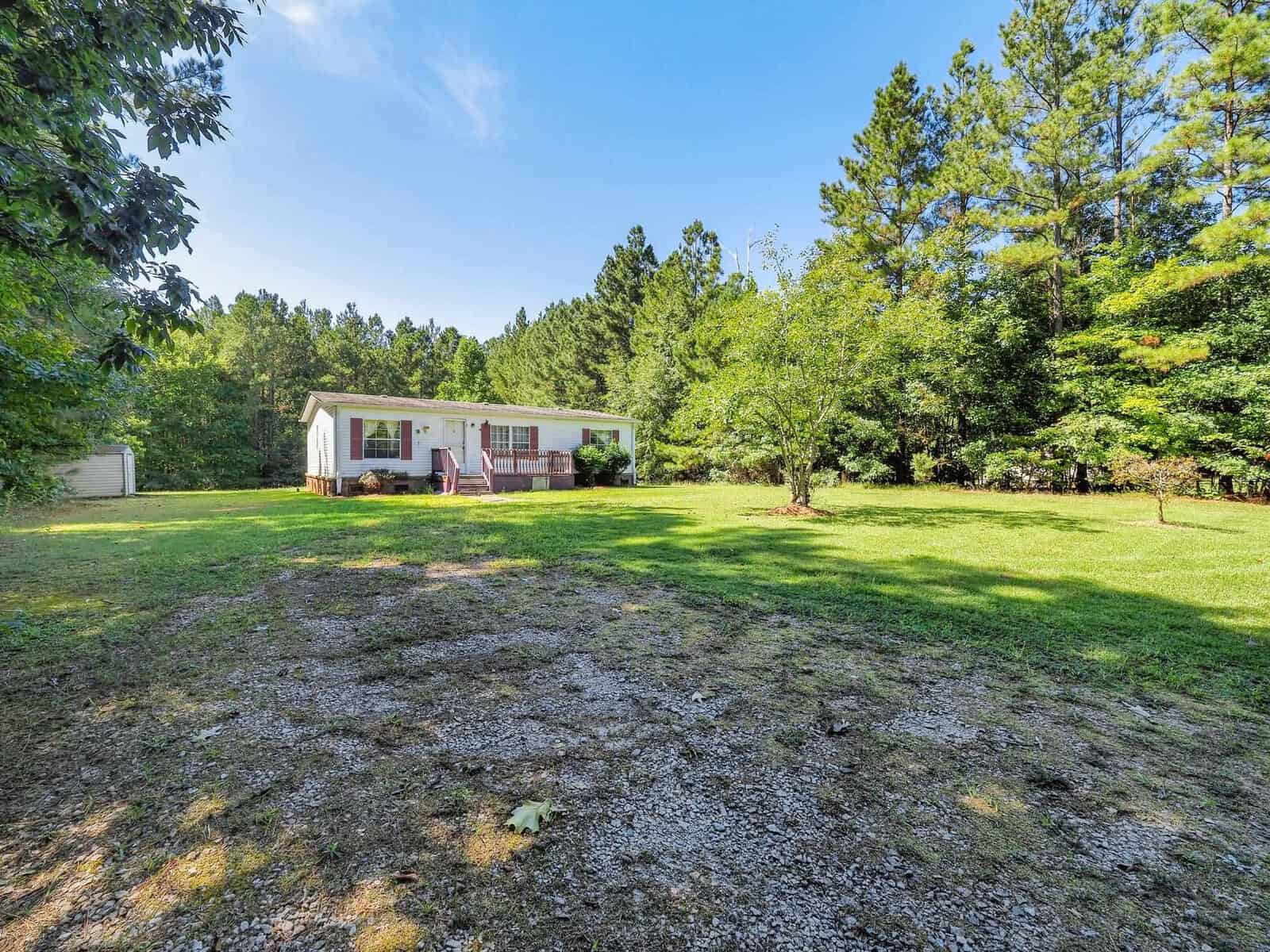 Small white house with a red door sits on a spacious, grassy lawn surrounded by trees. The sky is clear and blue, and a gravel path leads up to the house.