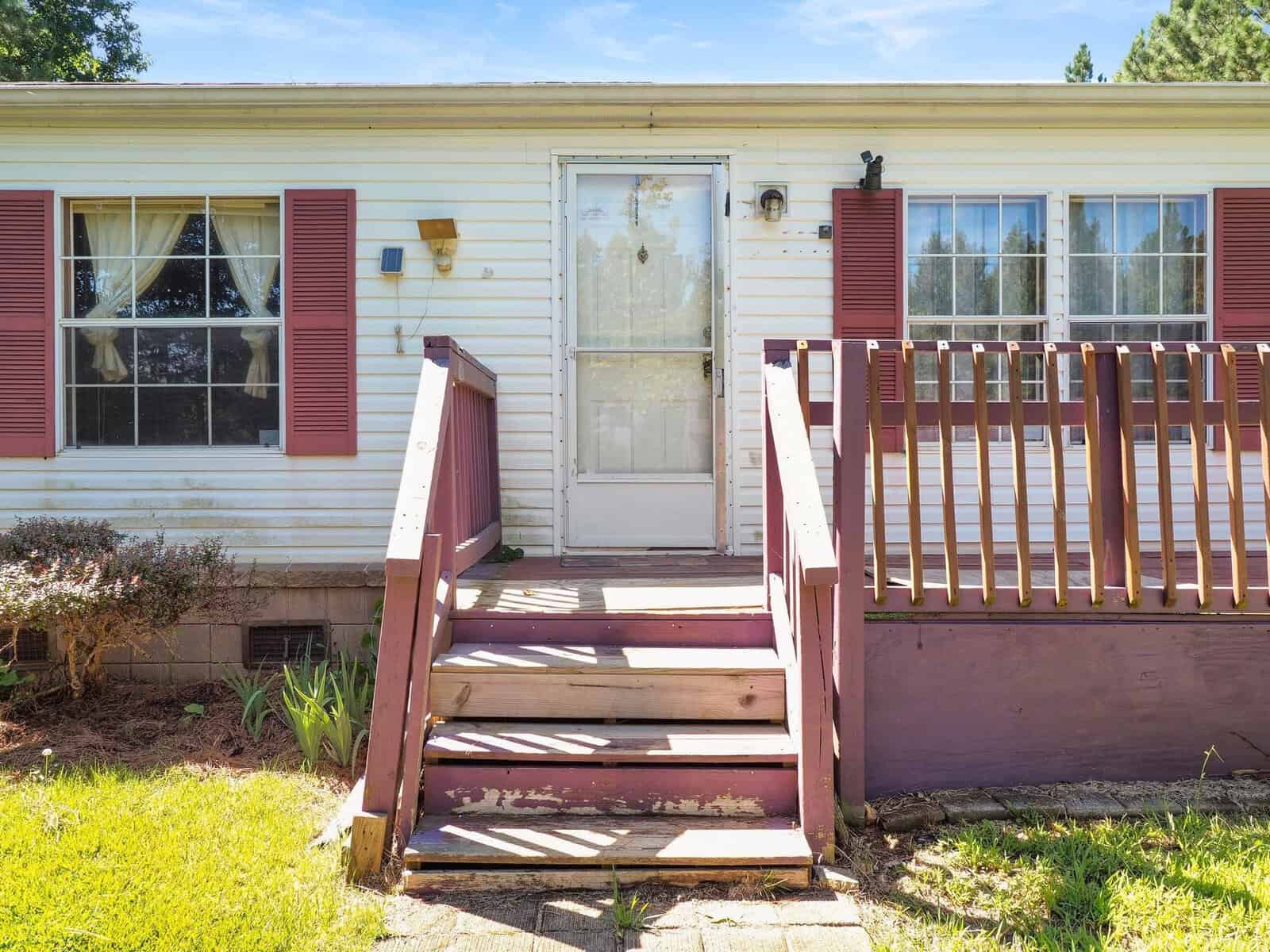 A small white house with red shutters and a front porch. The porch has stairs with red railings, leading to a screen door. There are two large windows with curtains on either side of the door. The house is surrounded by green grass and shrubs.